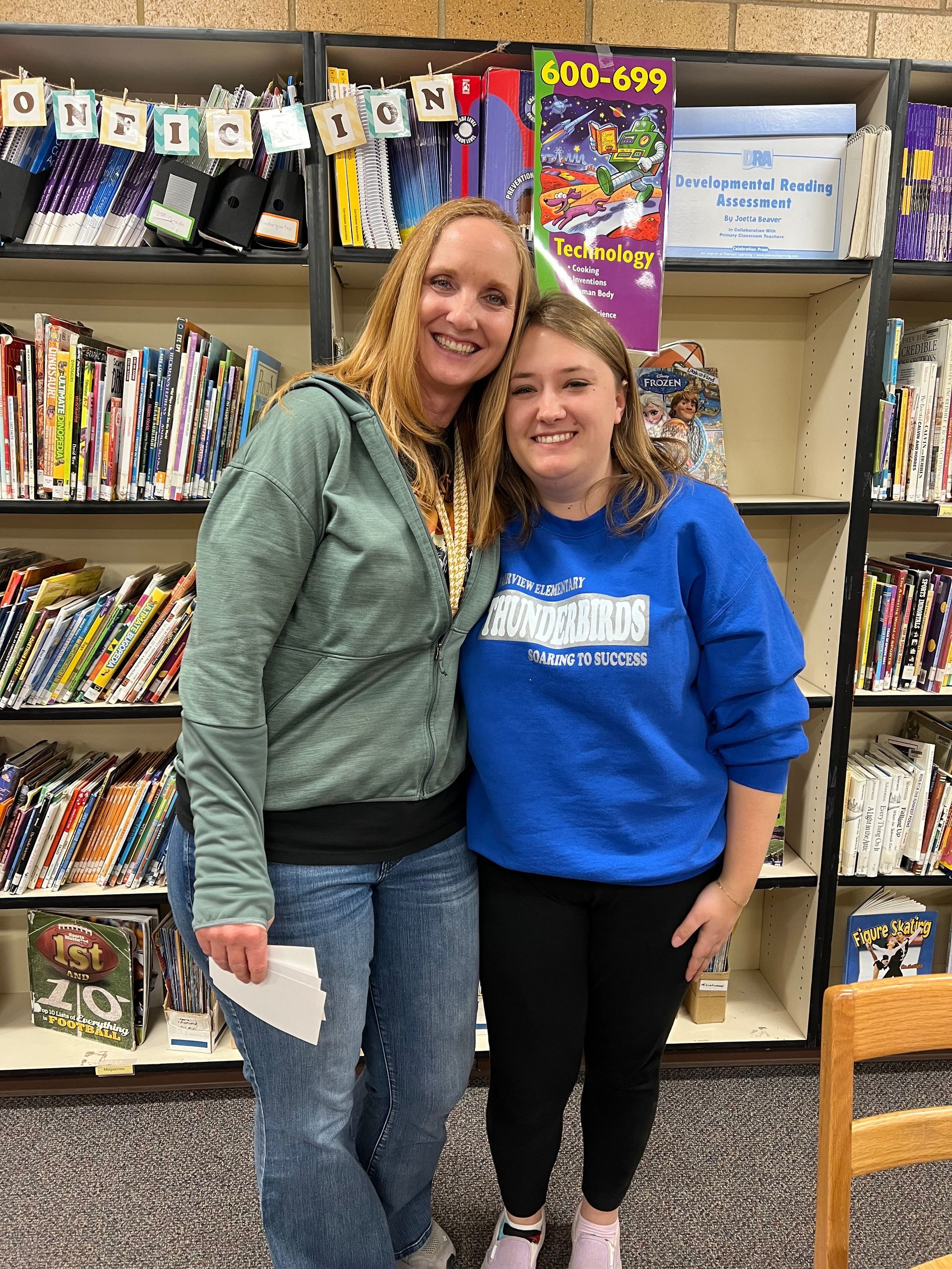 Two women are posing for a picture in front of a bookshelf.