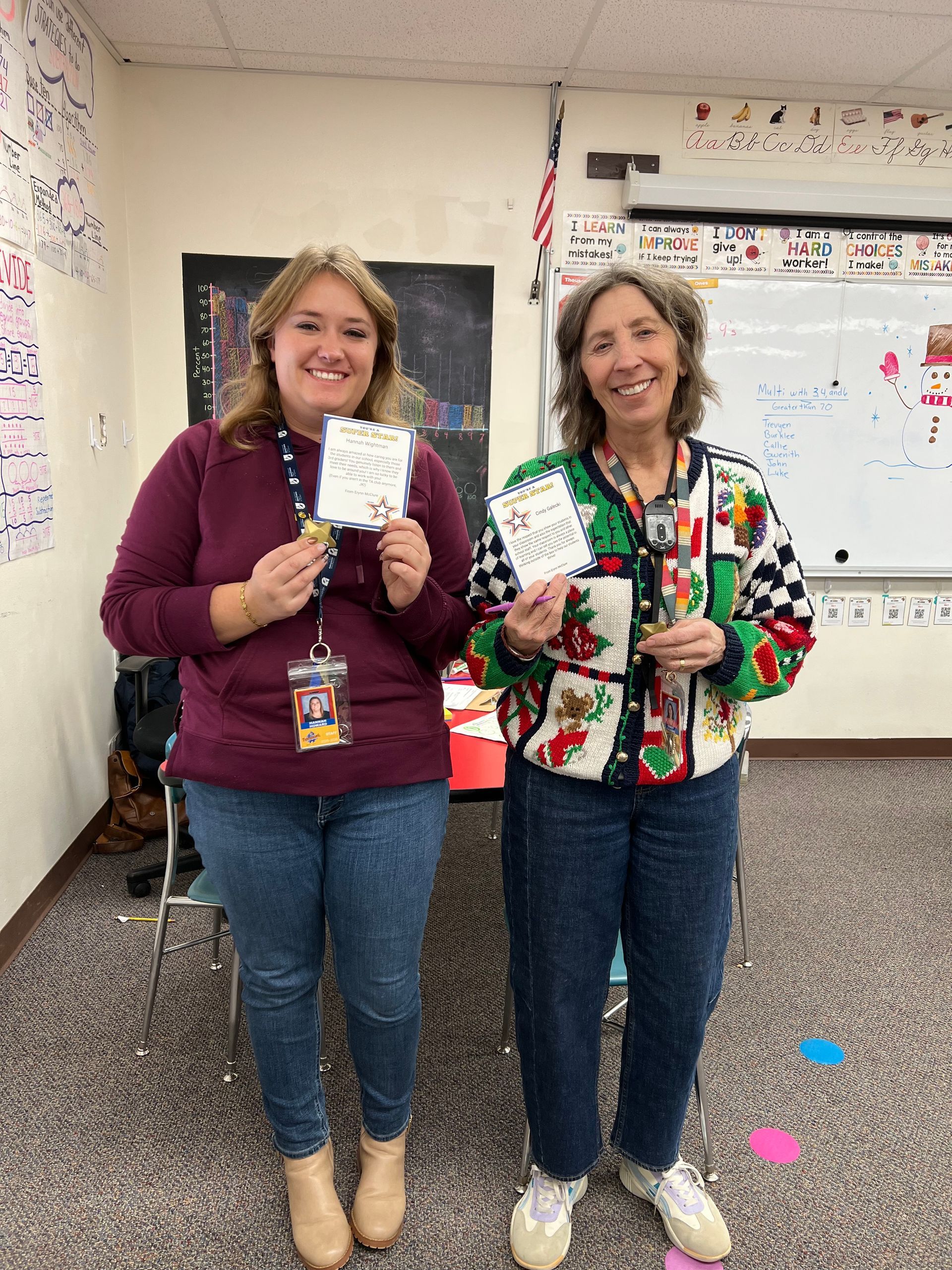 Two women holding cards, smiling in a classroom. One wears a maroon shirt and jeans; the other, a sweater and jeans.