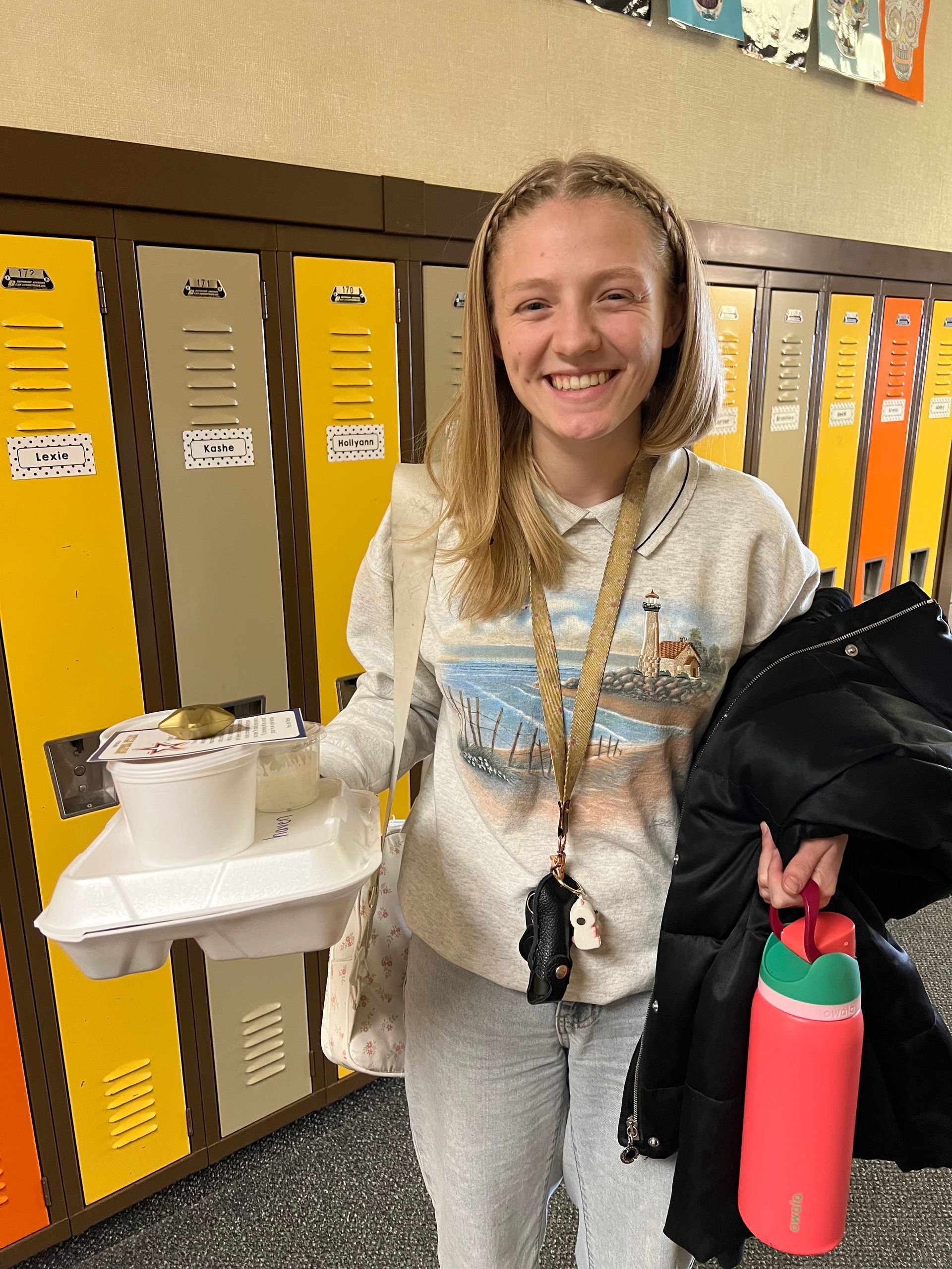 Woman holding food container, coat, and water bottle smiles in front of lockers.
