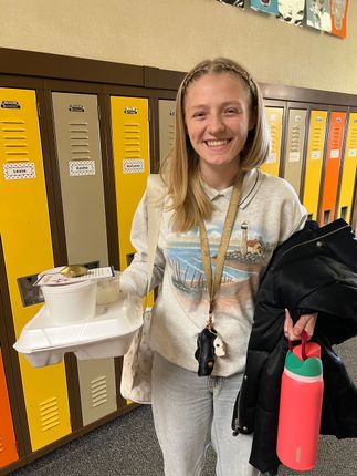 Woman holding a container of food, smiling, standing in front of yellow and brown lockers, holding jacket and water bottle.
