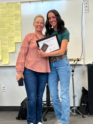 Two women stand in a classroom holding a framed certificate and smiling at the camera.