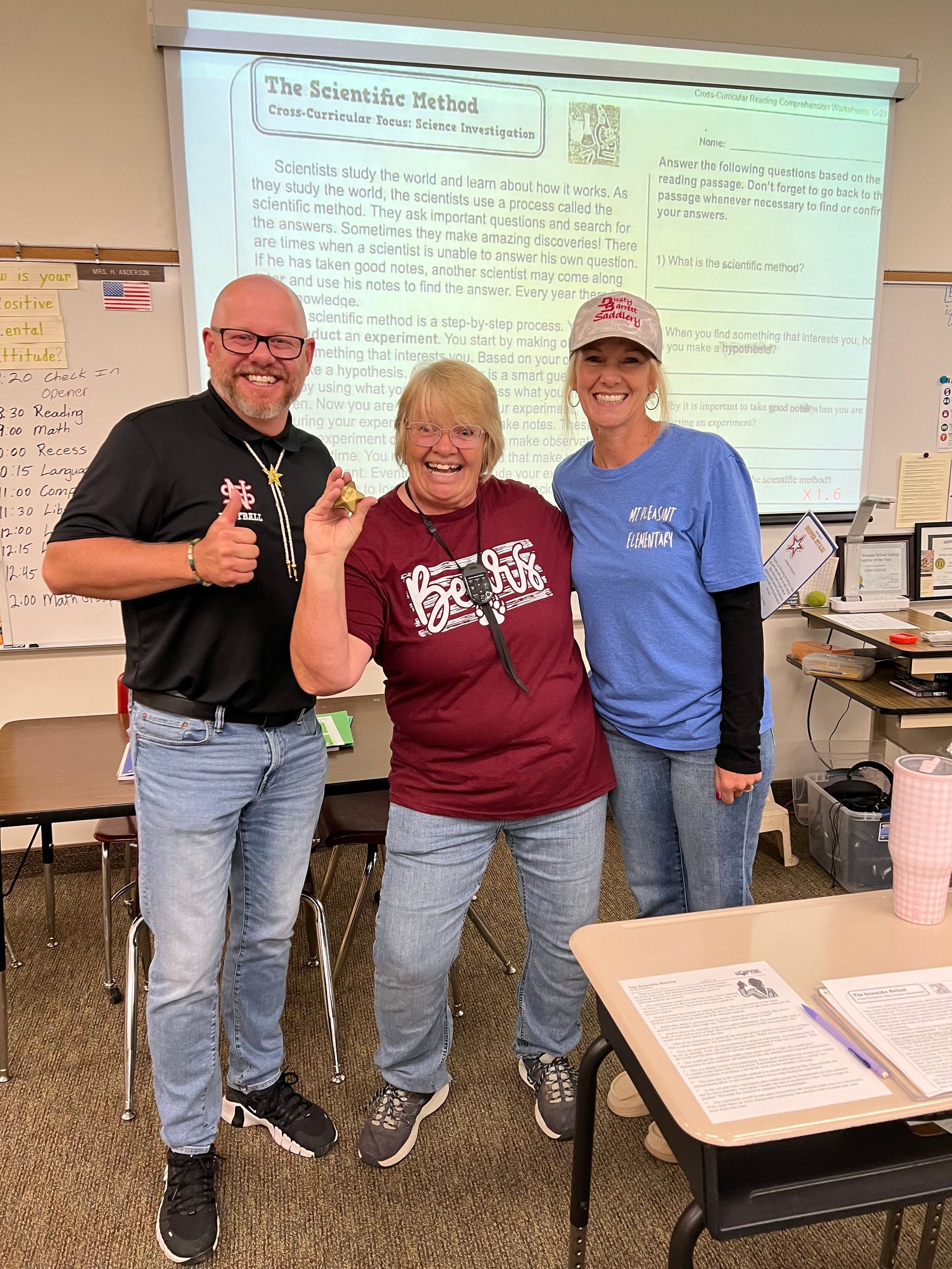 Three people in a classroom: a man with a thumbs up, a woman holding a medal, and a woman smiling.