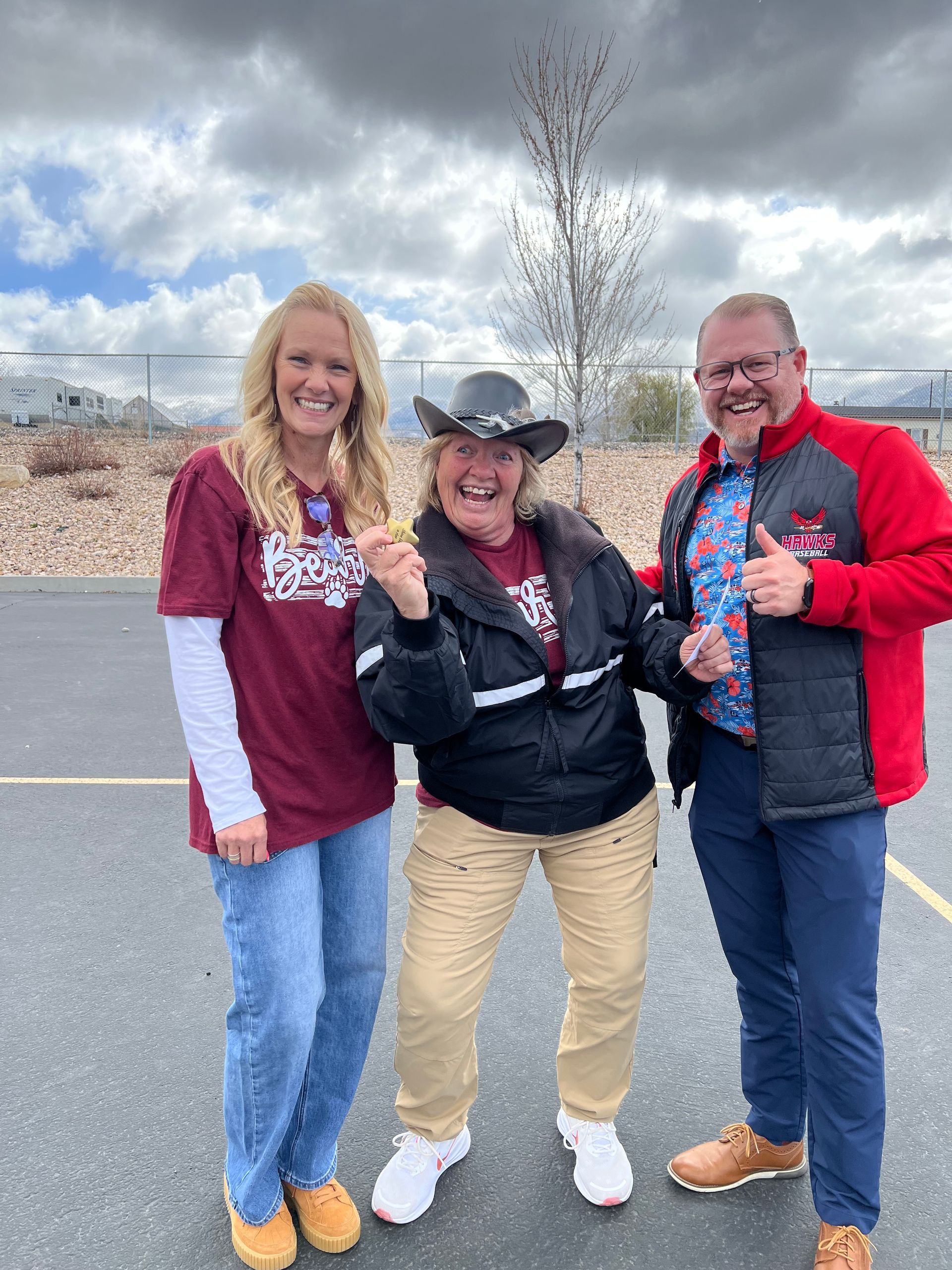 Three people smiling and posing outdoors on a road under a cloudy sky, wearing casual jackets and jeans.