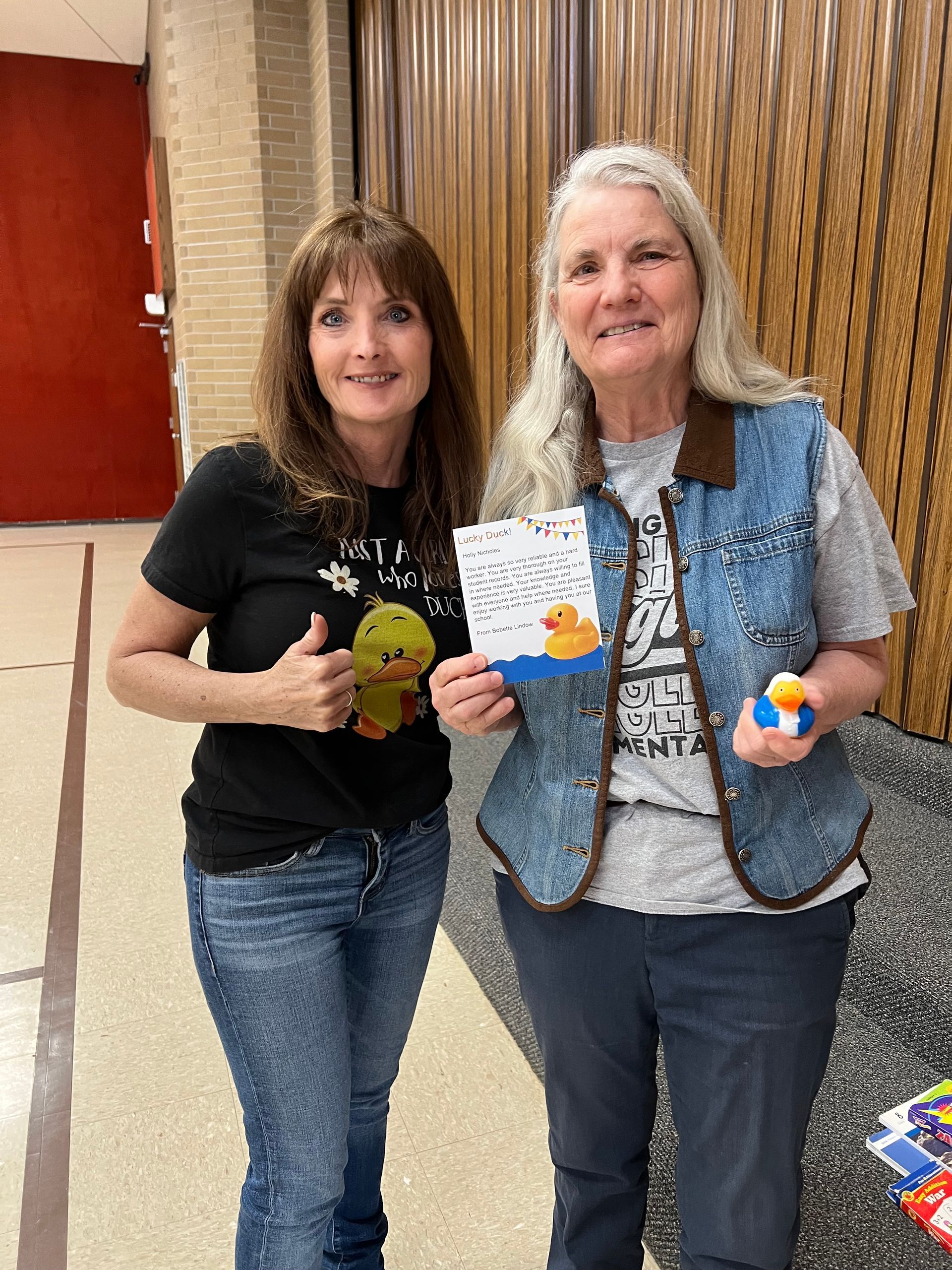 Two women are standing next to each other in a hallway holding a rubber duck.