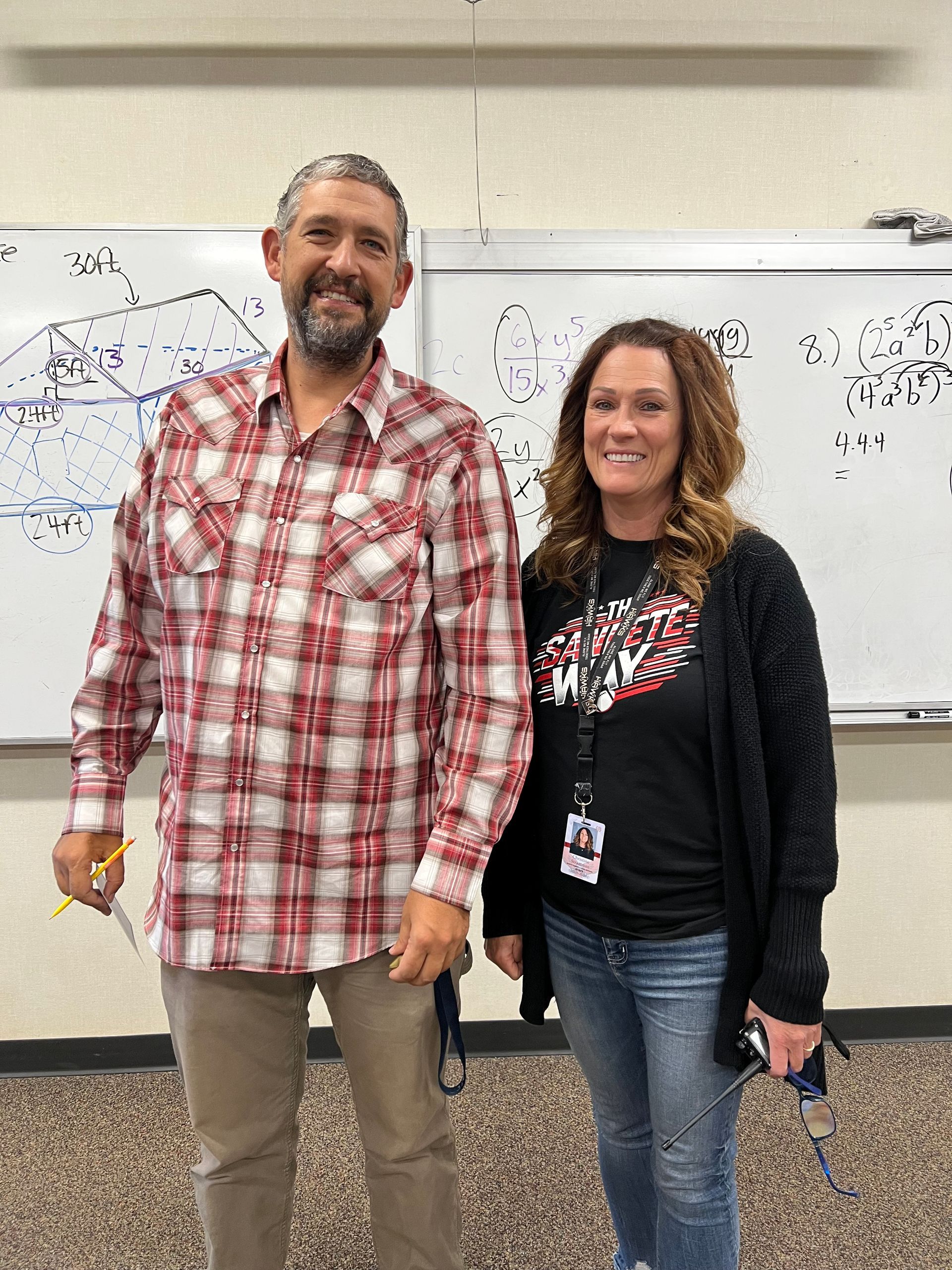 Two people standing in front of a whiteboard. Man in plaid shirt, woman in black shirt and cardigan. Classroom setting.