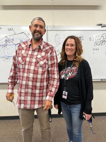 Two people standing in front of a whiteboard; the man is in a plaid shirt, and the woman is in a black shirt and cardigan.