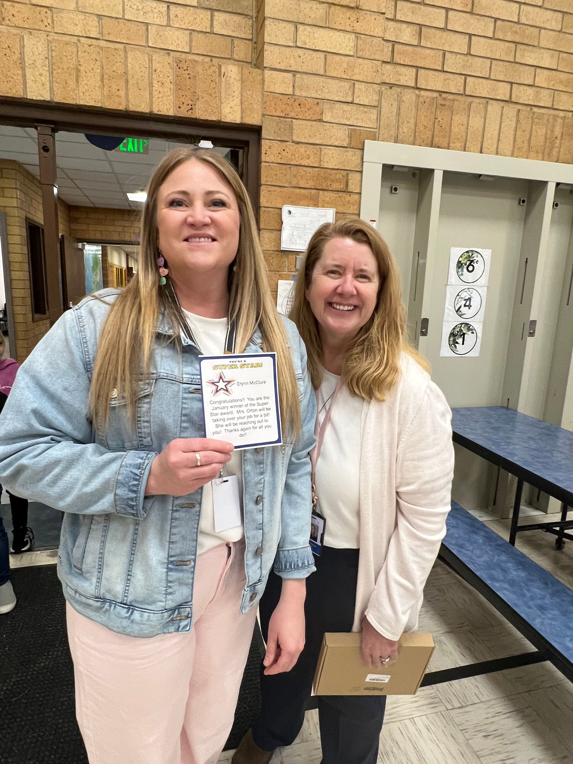 Two women smiling, one holding a card. Brick wall and lockers in the background.