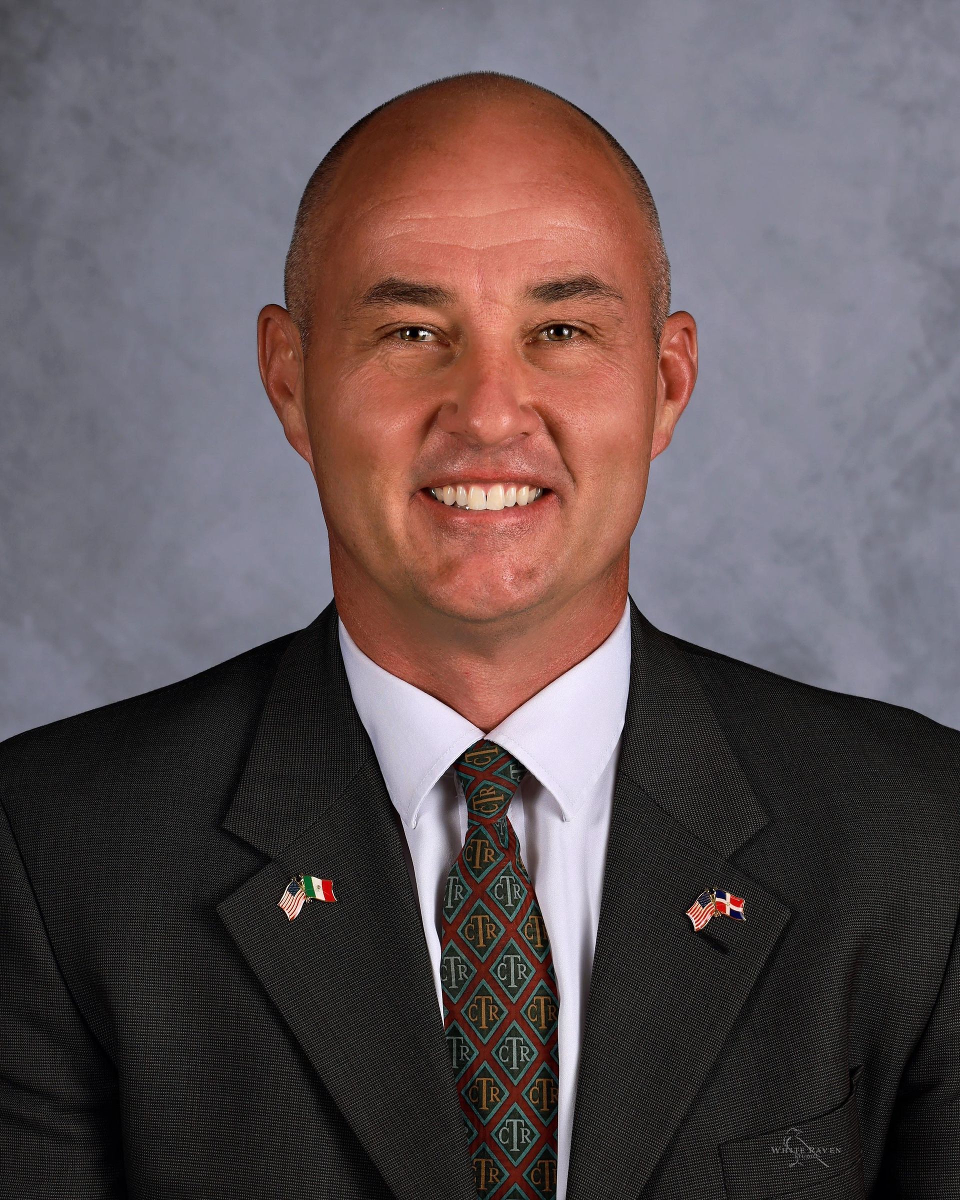Bald man in a suit smiles; small flags on lapel.