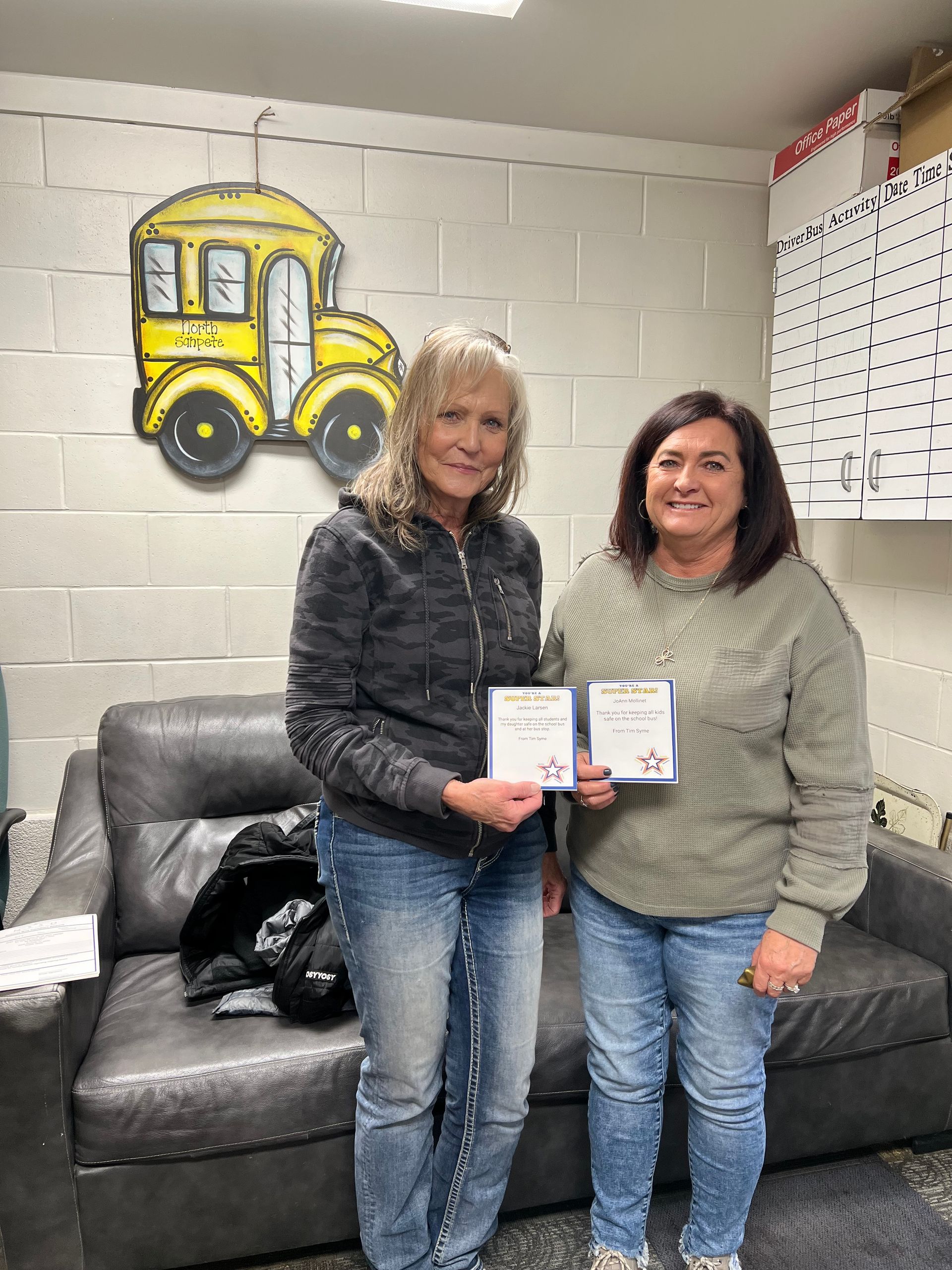 Two women holding certificates near a yellow school bus artwork. They are in a room with a gray couch and white brick wall.