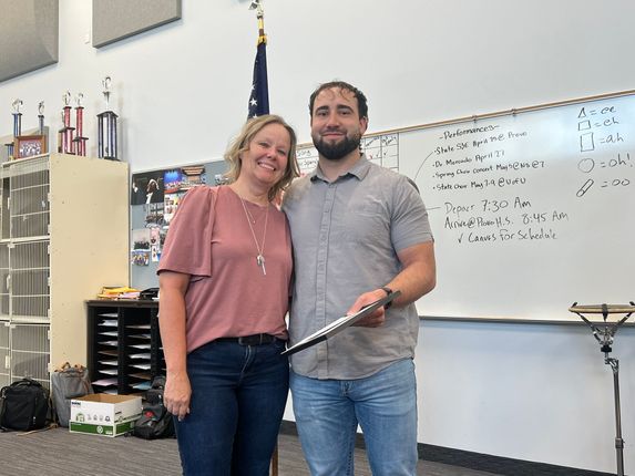 Two people posing in a music classroom, smiling and holding a certificate in front of a whiteboard