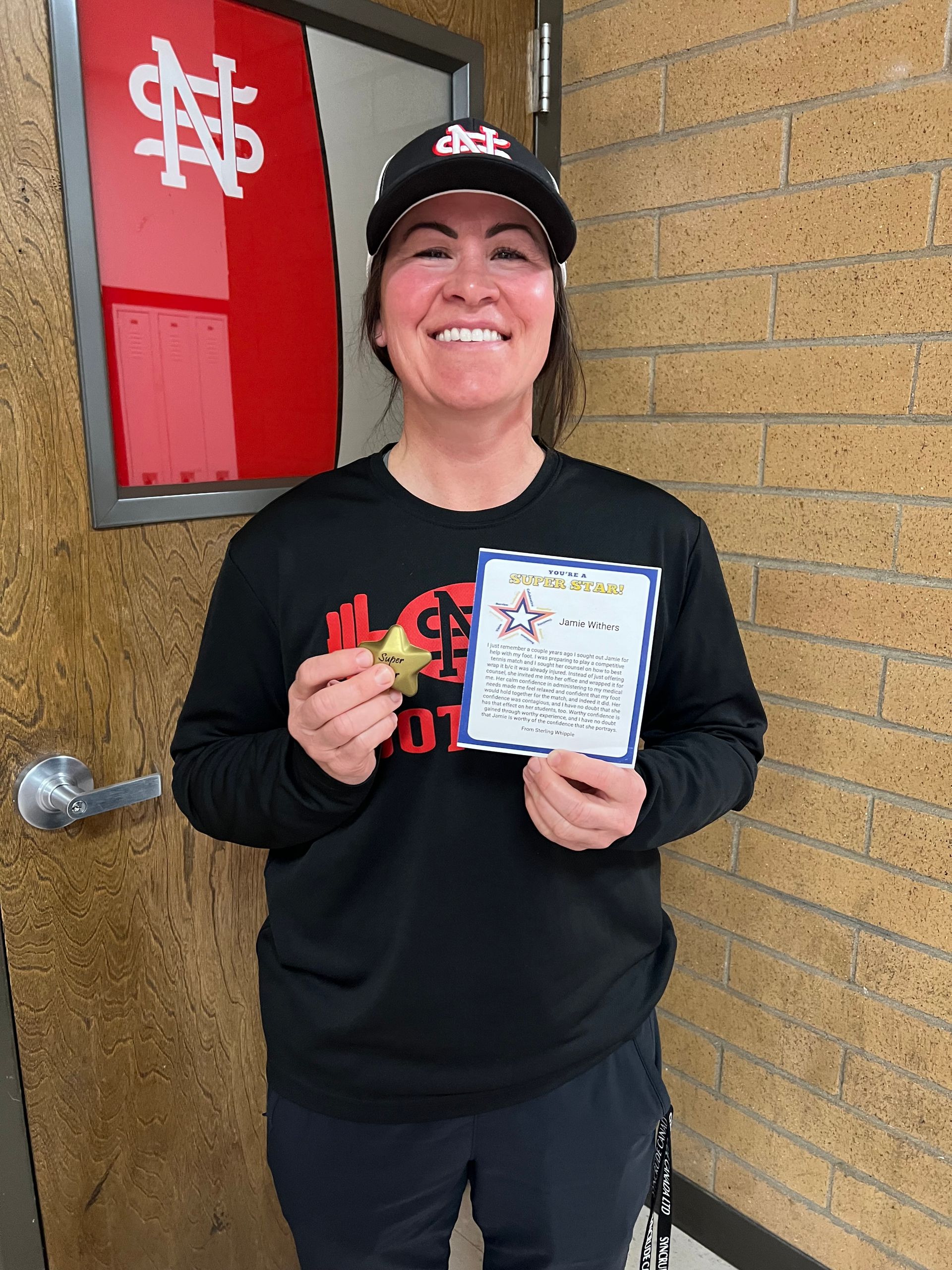 Smiling person wearing an athletic cap and black long-sleeve shirt holding a gold star and a certificate against a wall.