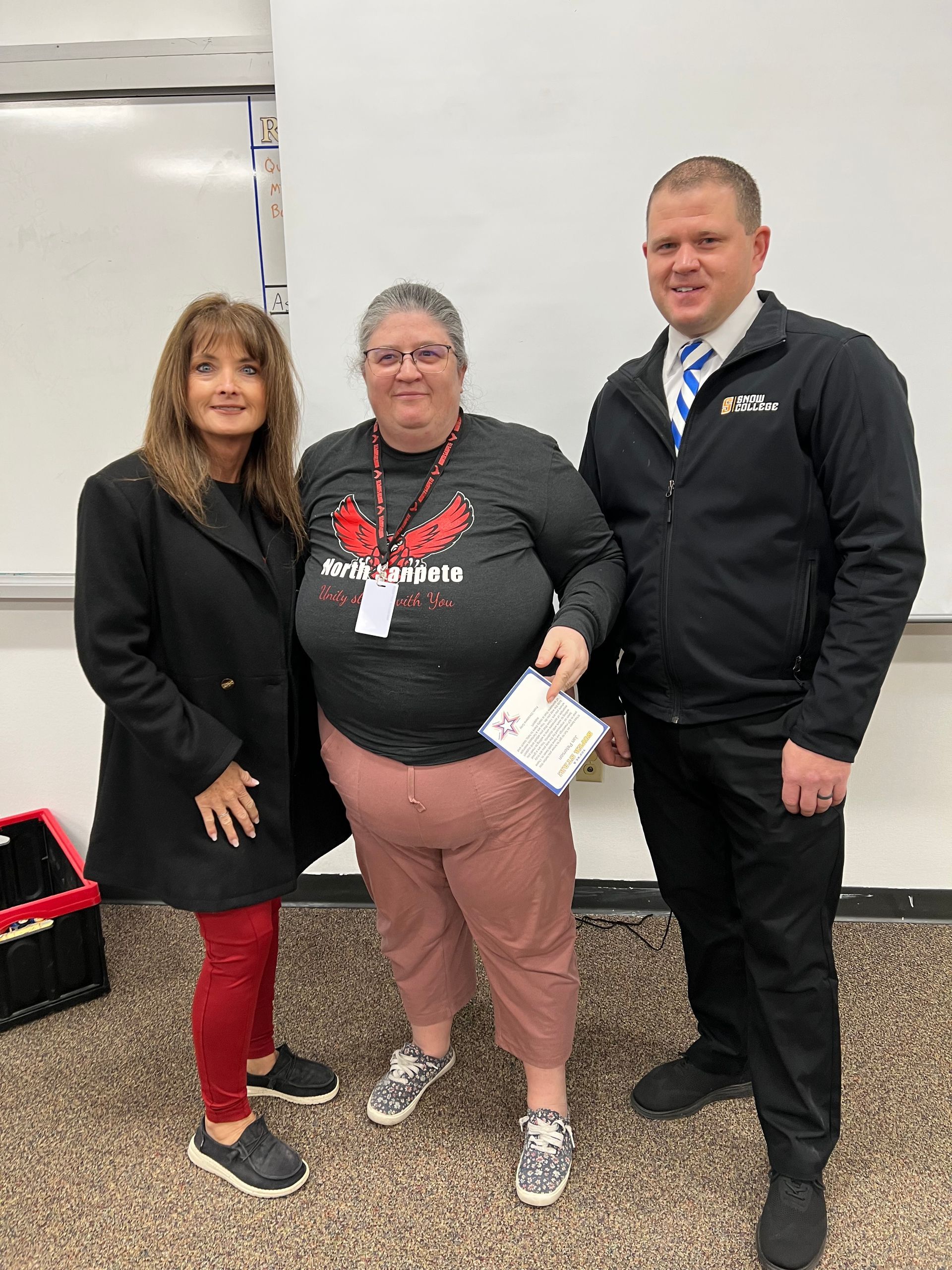 Three people pose indoors: woman in red pants, woman in grey shirt, man in jacket.