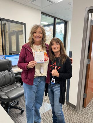 Two women pose in an office. One holds a card, the other gives a thumbs-up.