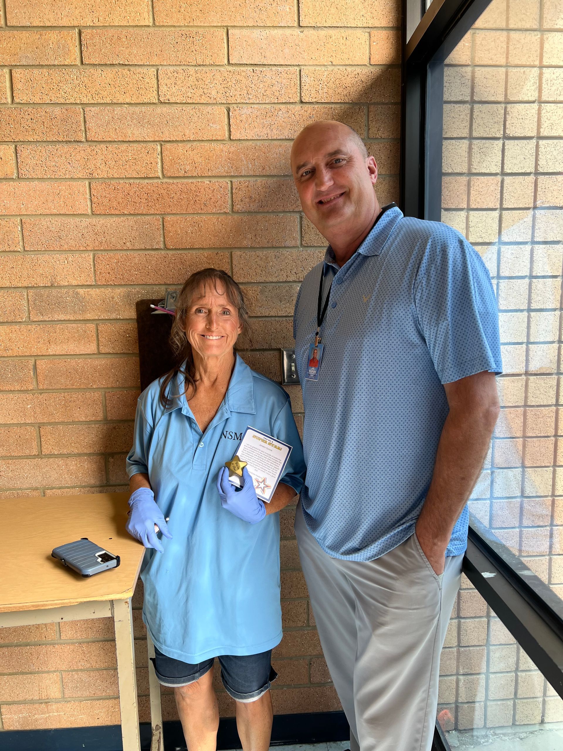 Two people stand near a brick wall and a window. Woman in blue scrubs and man in a blue polo shirt.