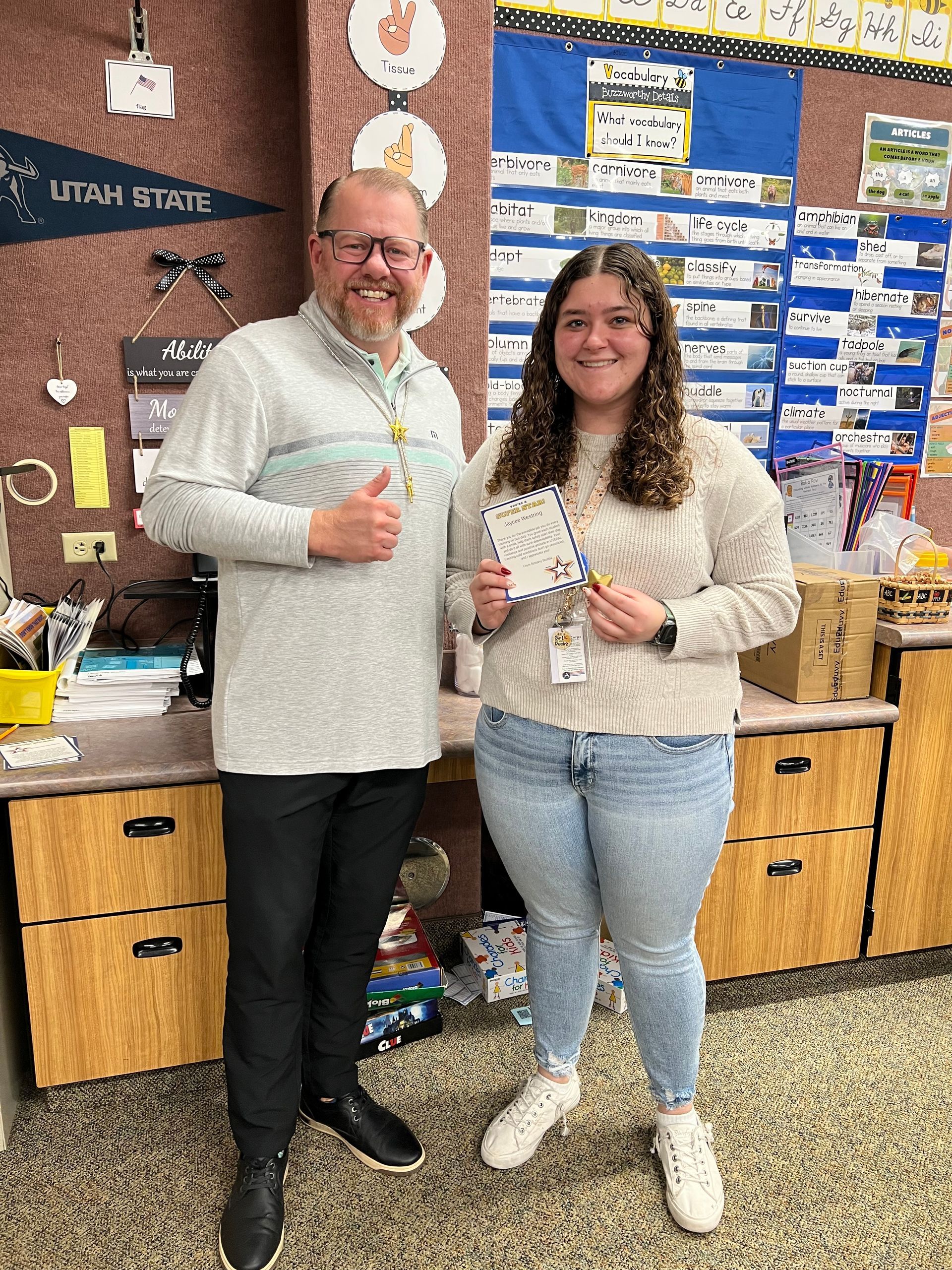A man and woman in a classroom. The man gives a thumbs up; the woman holds a pencil and paper.