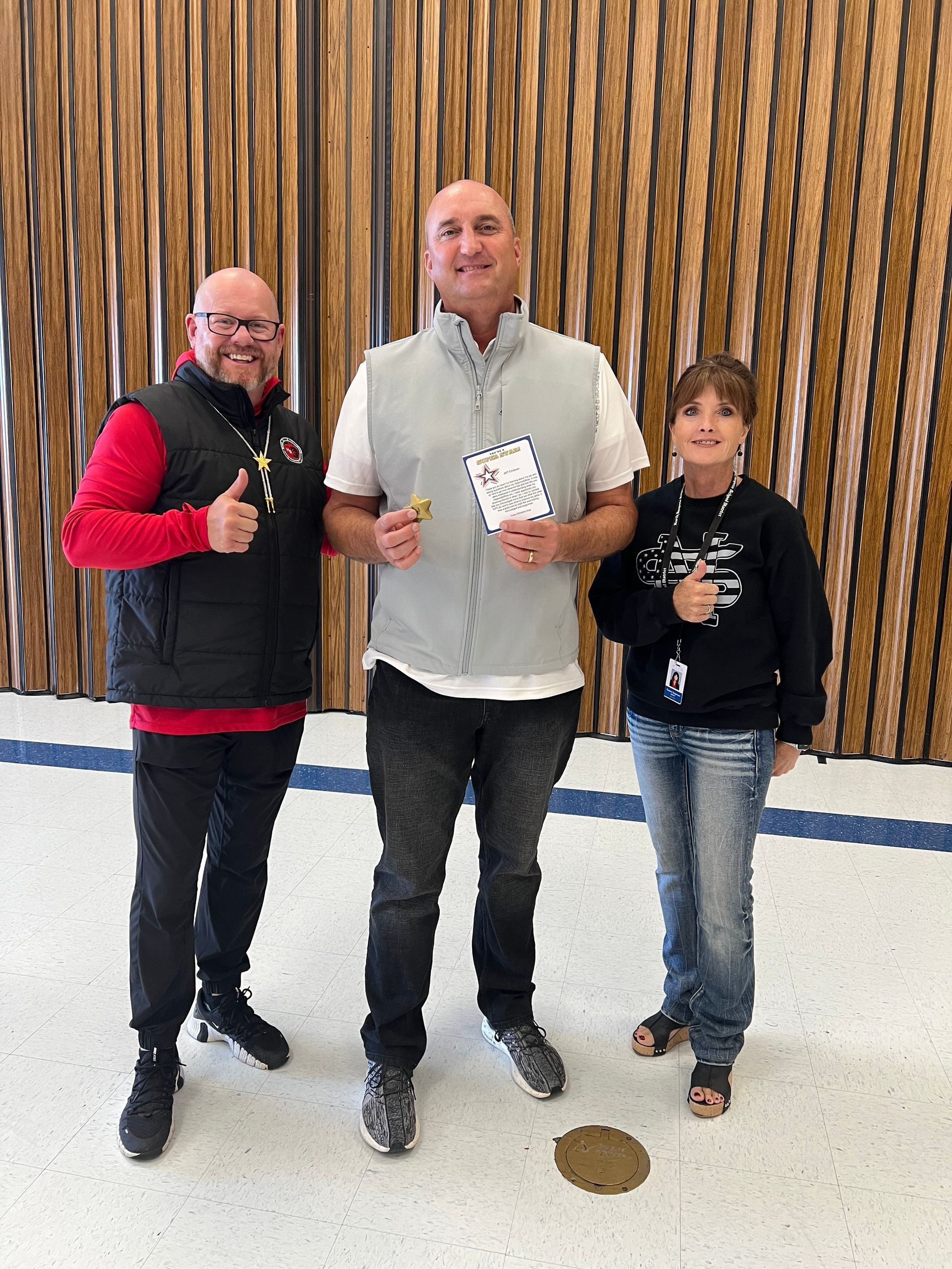 Three people smiling and giving thumbs-up: man in gray vest, woman in black sweatshirt, and man in red vest, indoors.