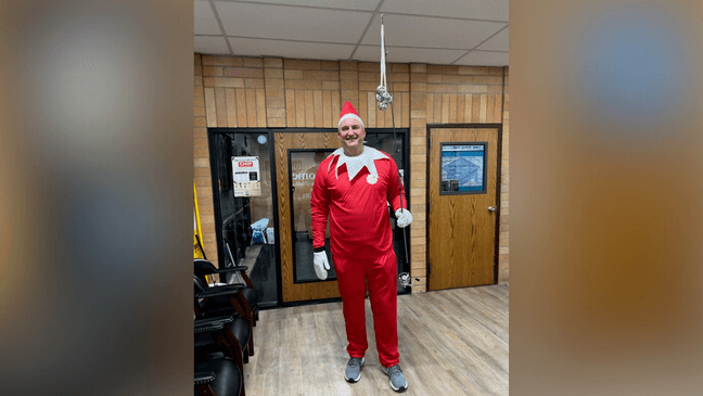 A man in a red elf on the shelf costume is standing in a room.