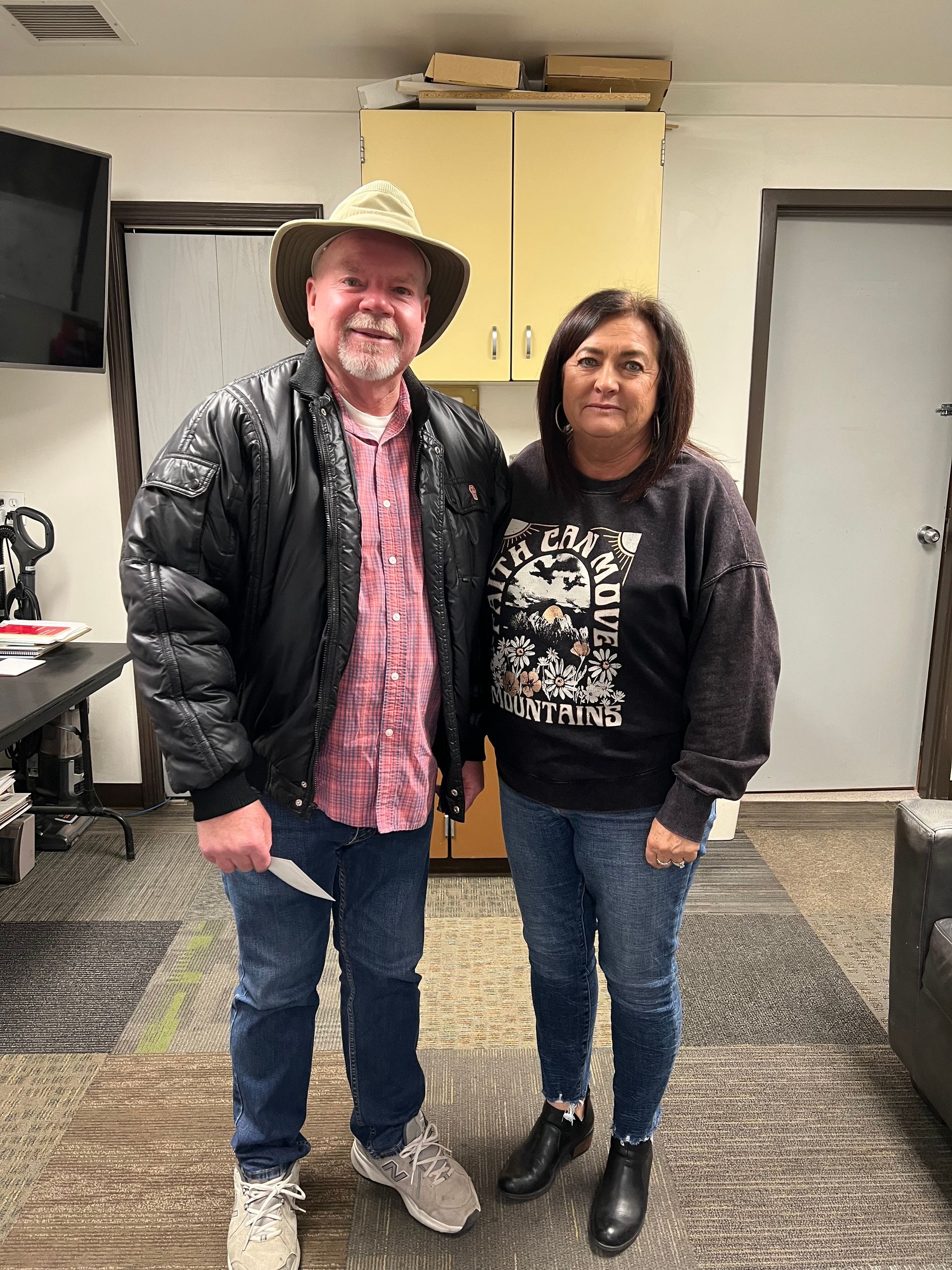 Man in jacket and hat stands next to a woman in a sweatshirt and jeans, indoors.