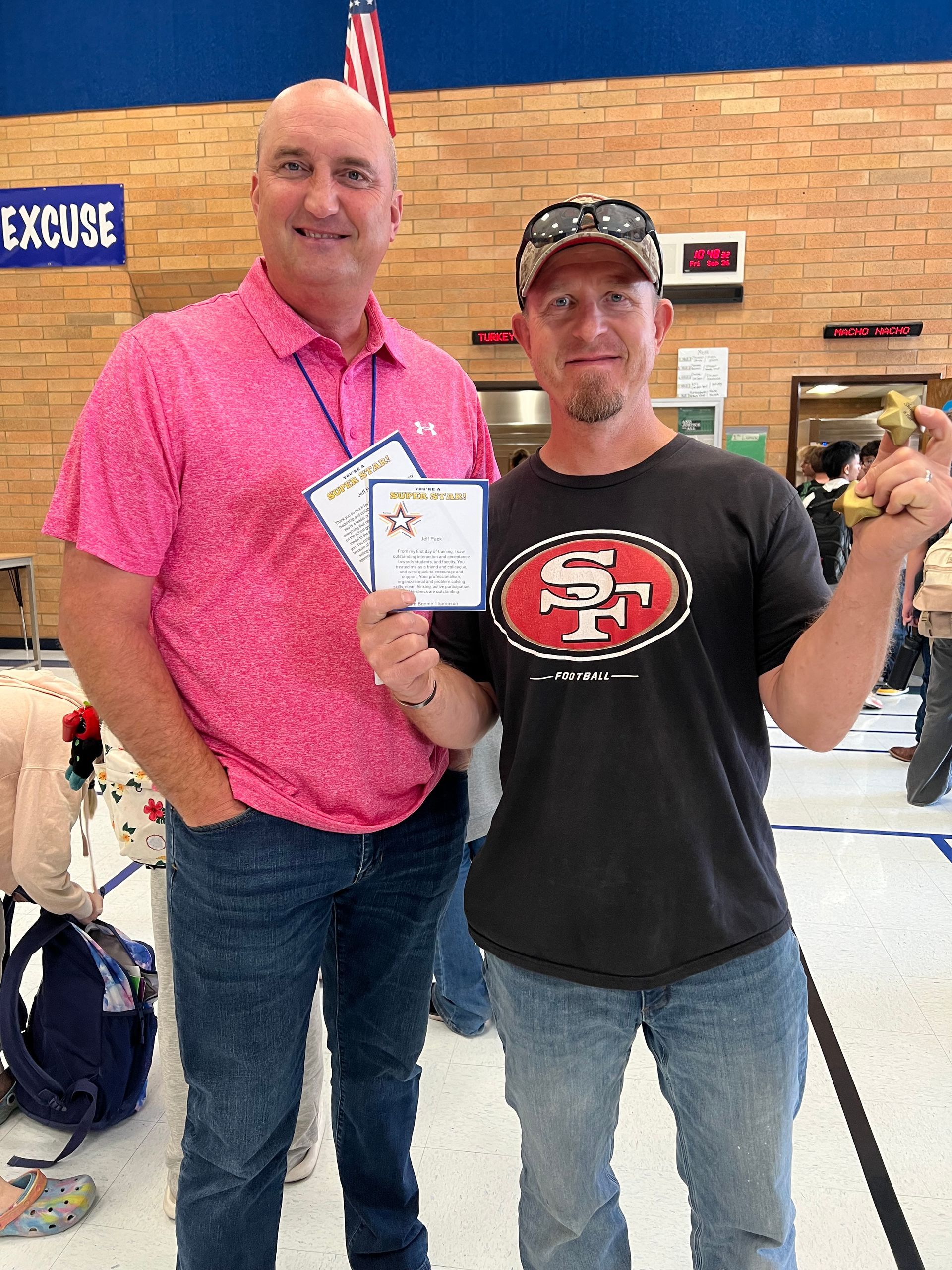 Two men smiling, one in pink shirt, the other in a 49ers shirt, holding certificates and a trophy in a school hallway.