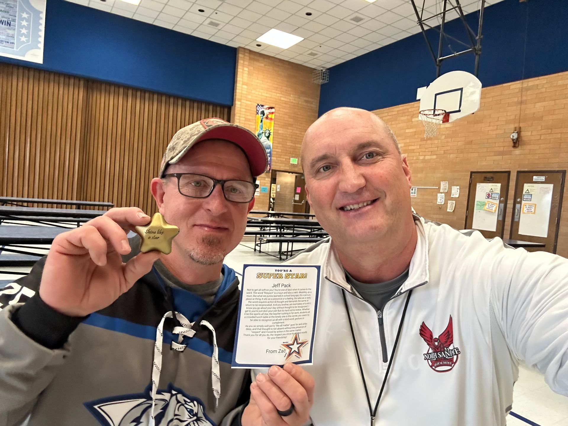 Two men in a school cafeteria; one holds a star award and a certificate. Blue and tan walls.
