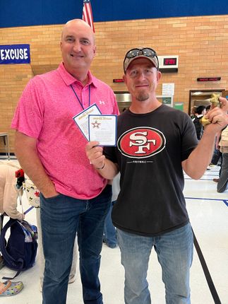 Two men pose, one in pink shirt and one in 49ers shirt holding a trophy and awards.