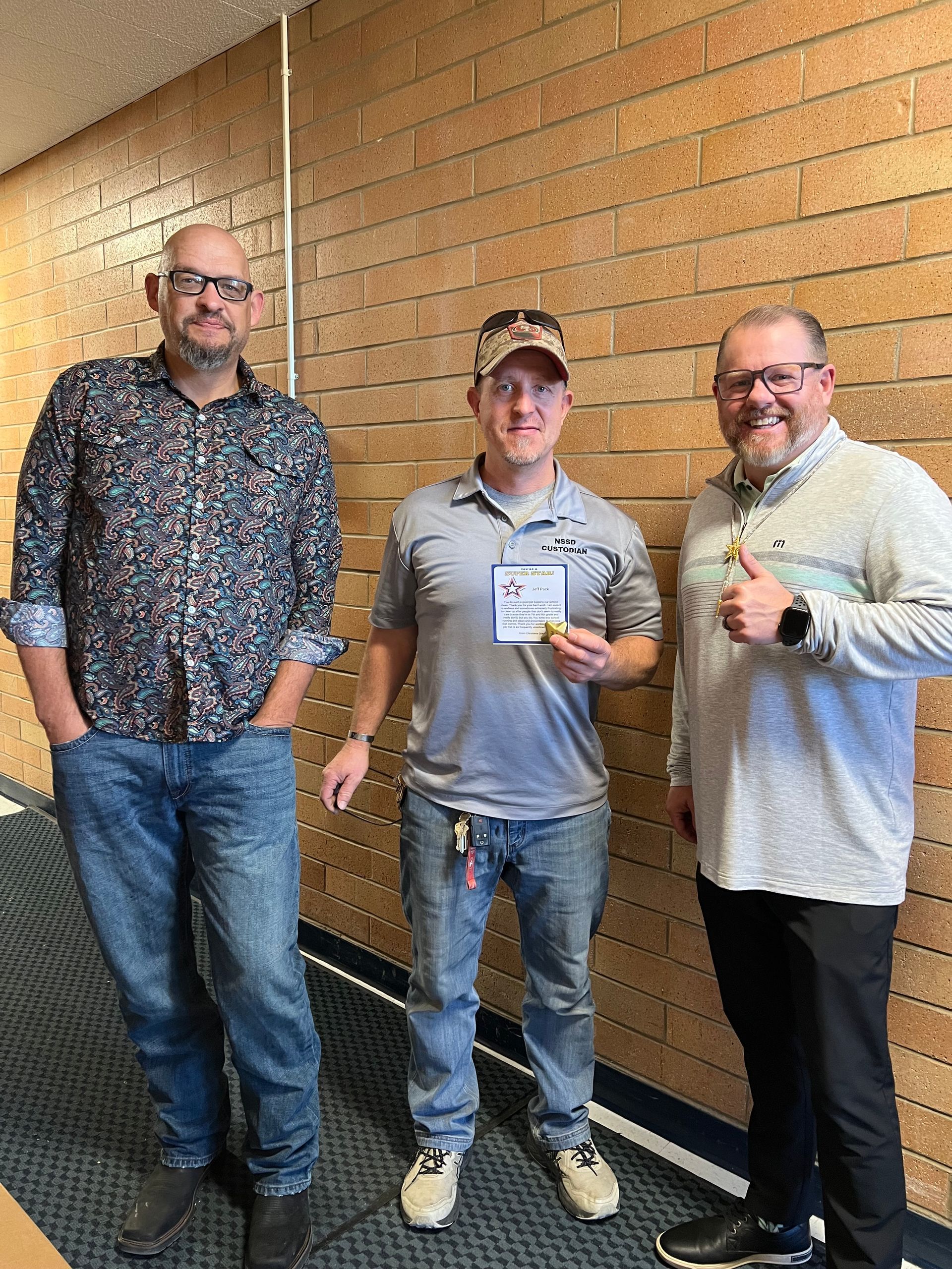 Three men pose in a hallway. The center man holds a name tag. One man gives a thumbs-up.