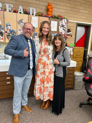 Three people smiling, posing together indoors. Woman in floral dress in the middle, others in business attire.