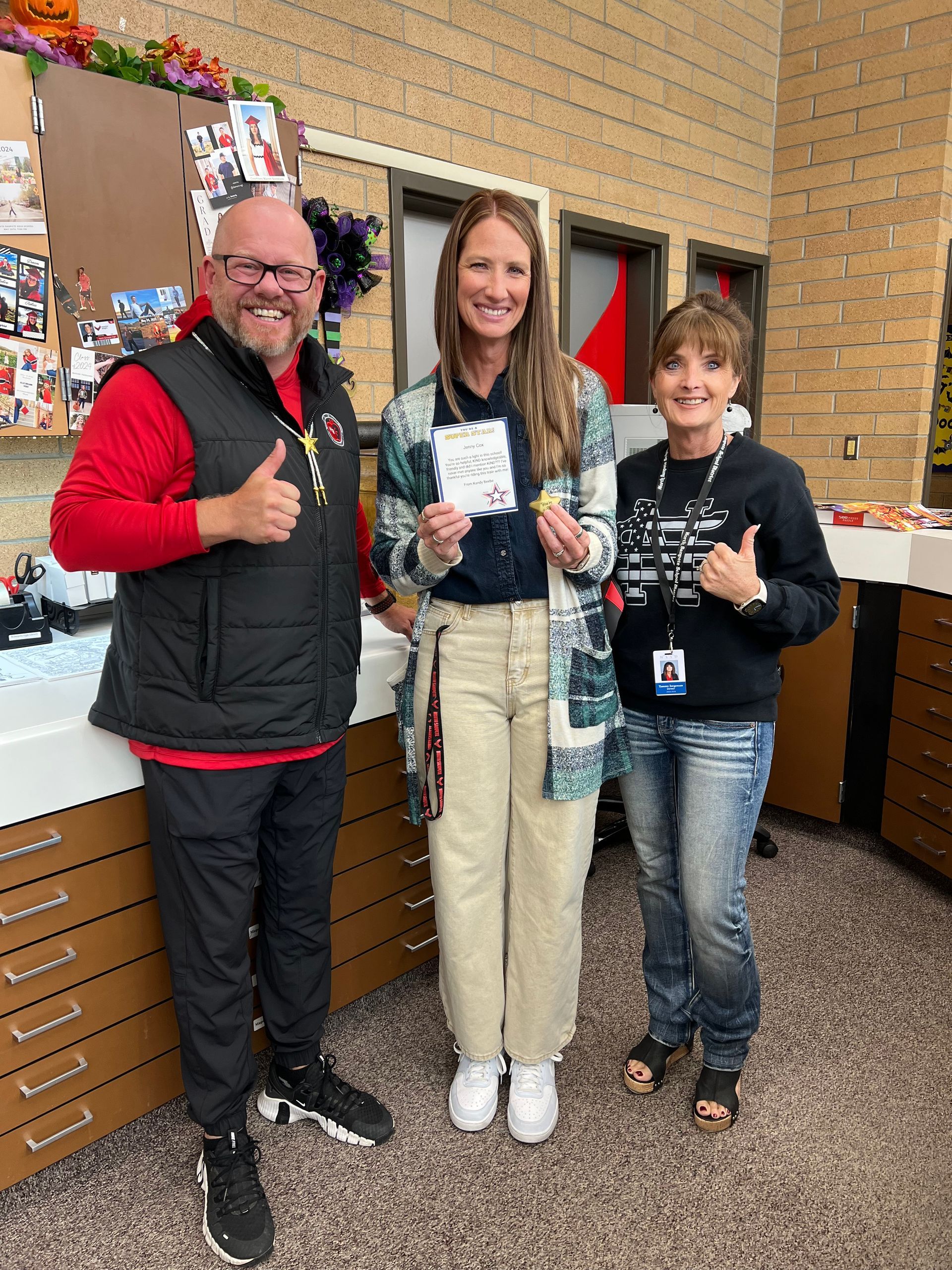 Three people standing together, smiling and giving thumbs up. A man on the left wears a red shirt and black vest. Woman holds paper.