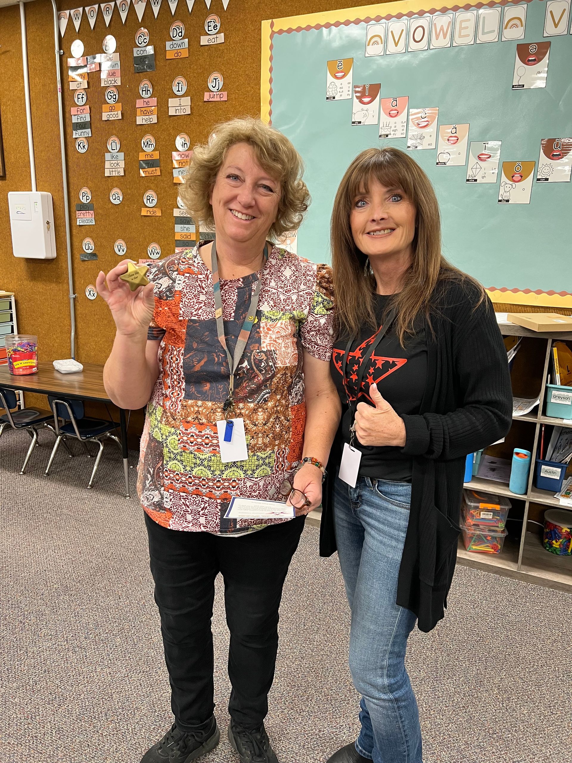 Two women in a classroom, one holding object, both smiling. Background: bulletin boards, desks, and shelves.