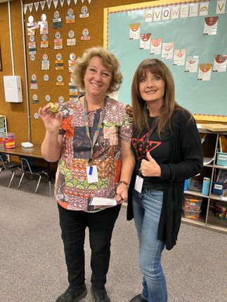 Two women in a classroom, one holding object, both smiling, with decorations in background.