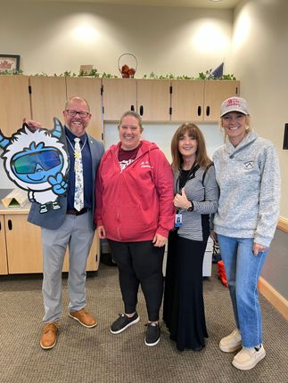 Four people smiling indoors, holding a yeti cutout, possibly a school setting.