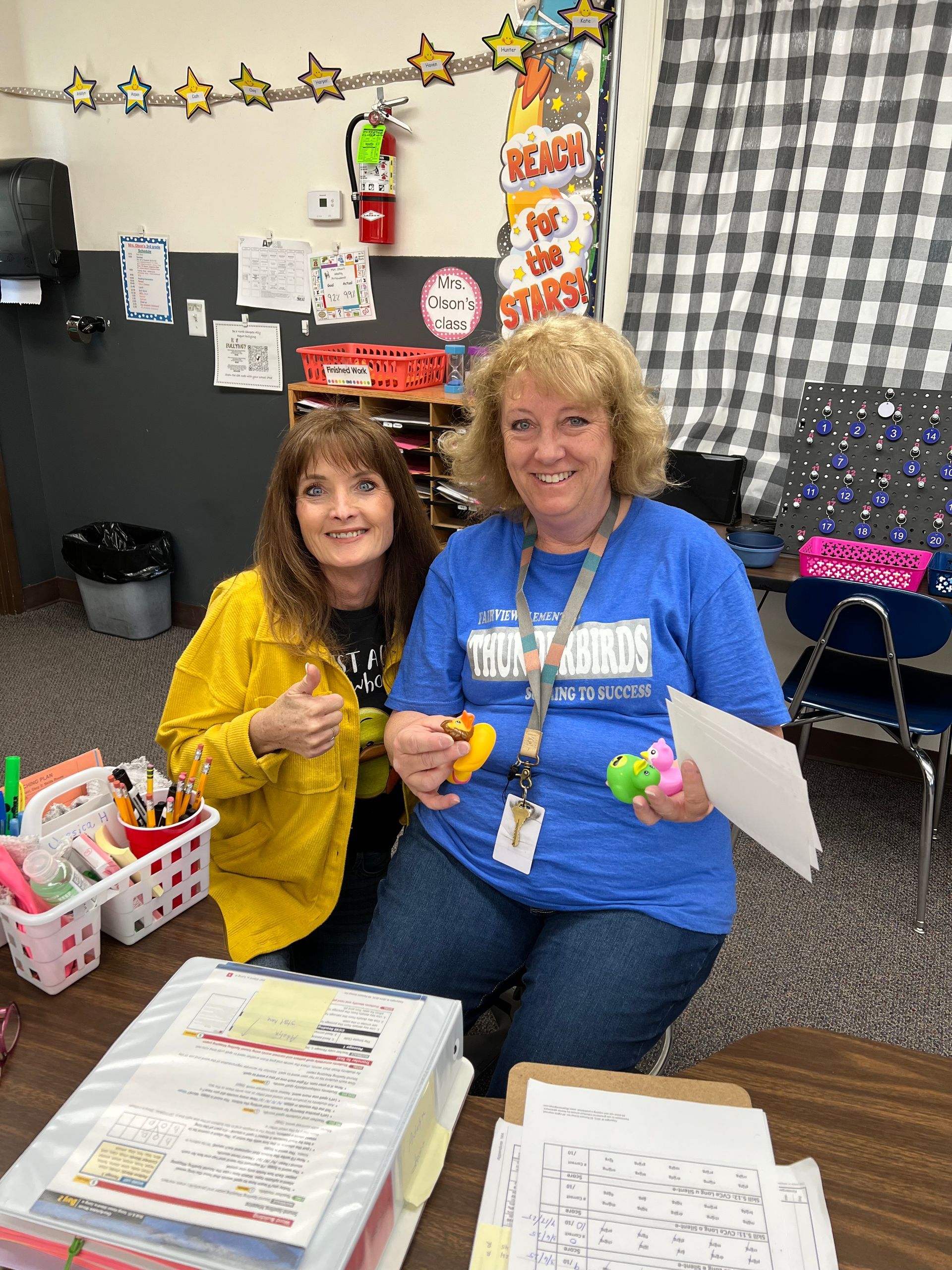 Two women are sitting at a desk in a classroom holding toys.