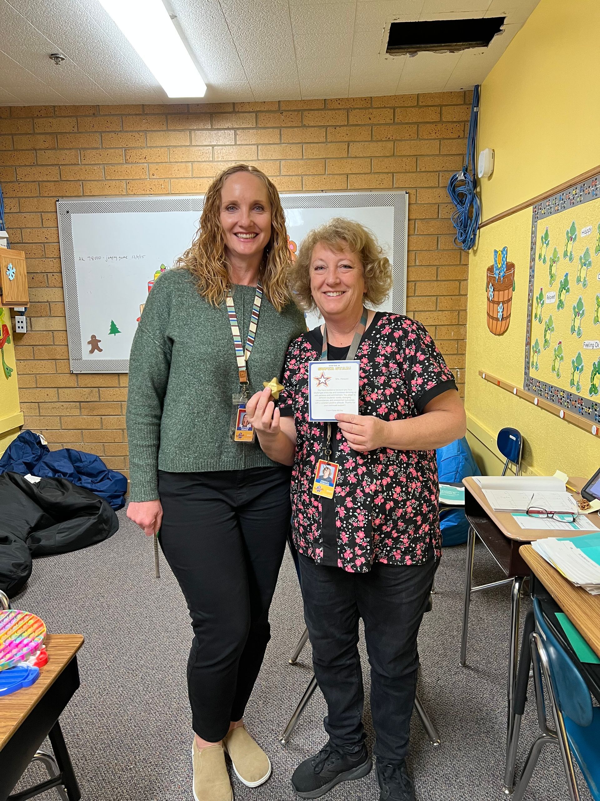Two women in a classroom; one holds a card. They smile, posing next to a whiteboard and desk.