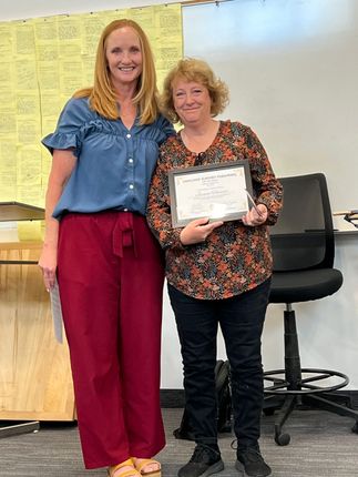 Two women smiling on stage, one holding a certificate, in front of a classroom chair and board.