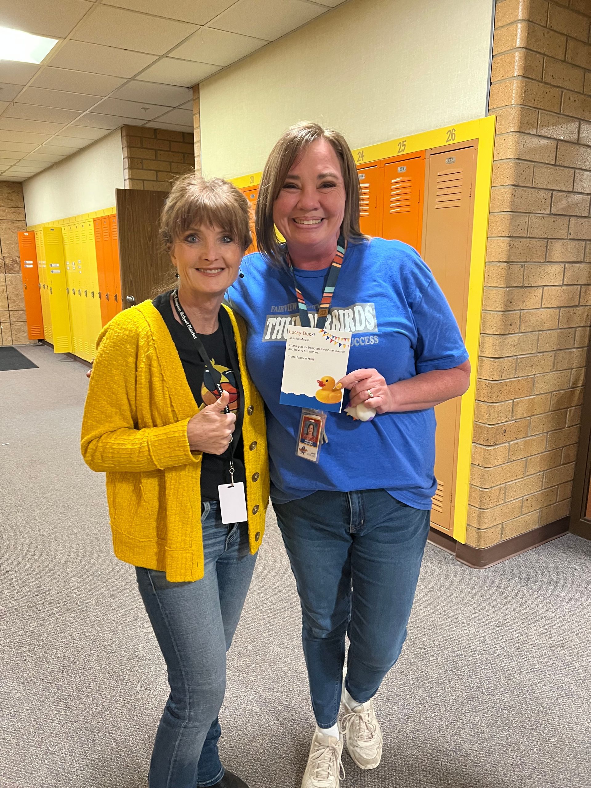 Two women are standing next to each other in a hallway in front of lockers.
