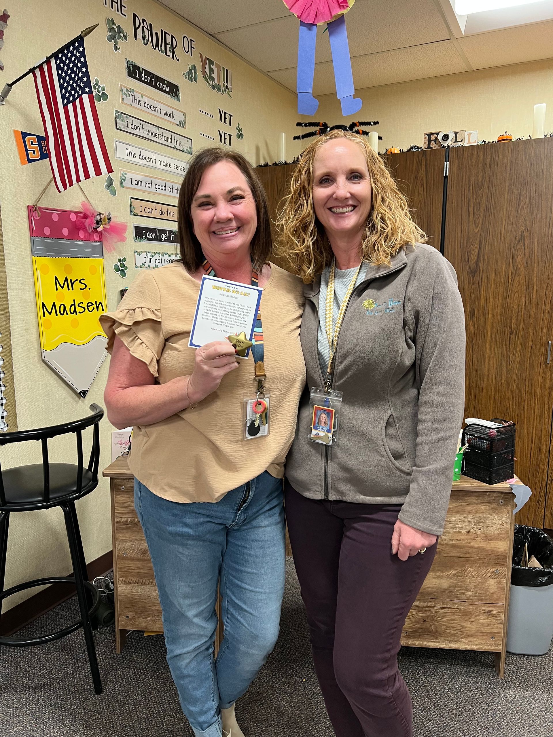 Two women smiling, holding a card, standing in a decorated classroom with an American flag.