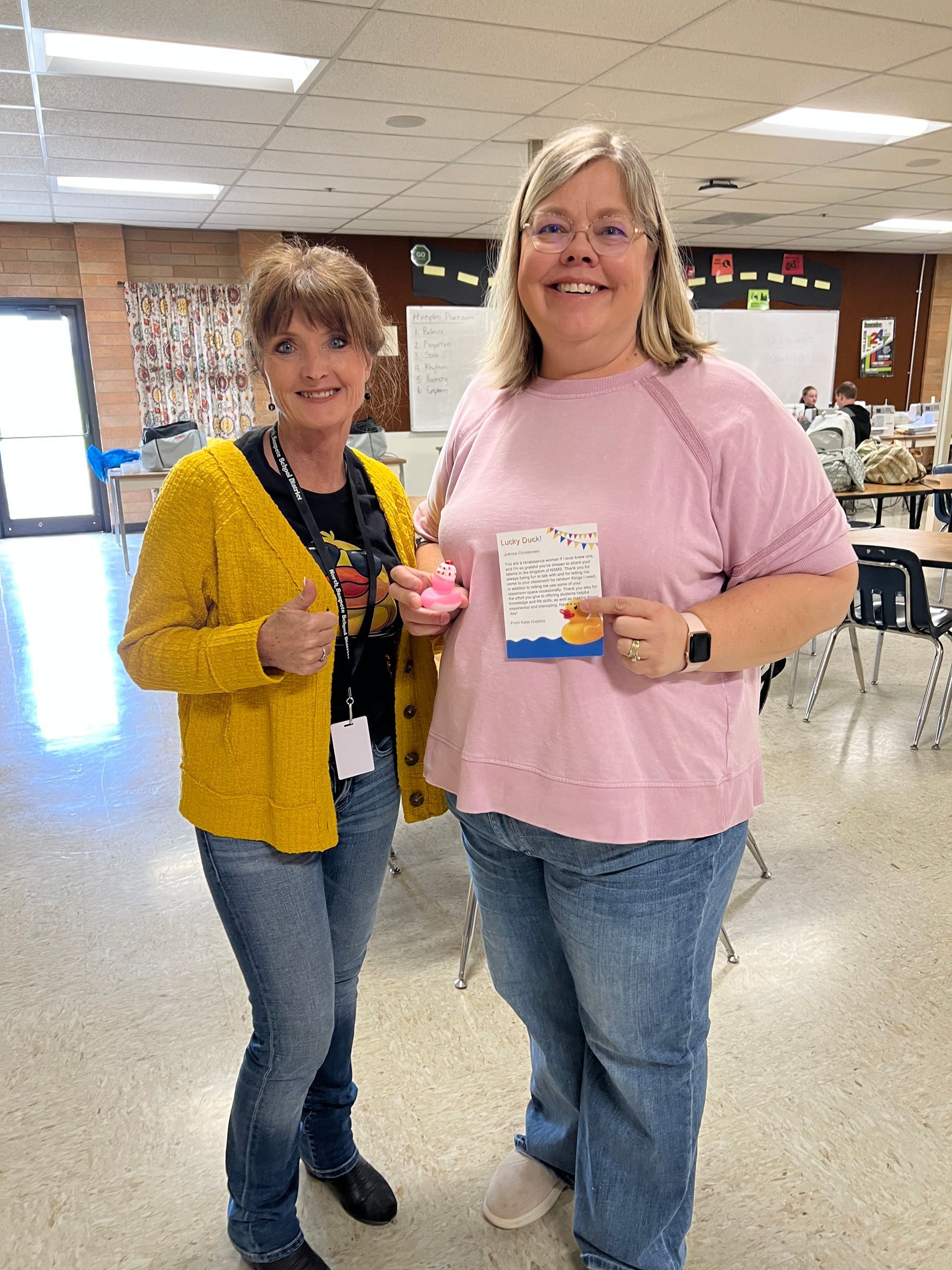 Two women are standing next to each other in a classroom holding donuts.