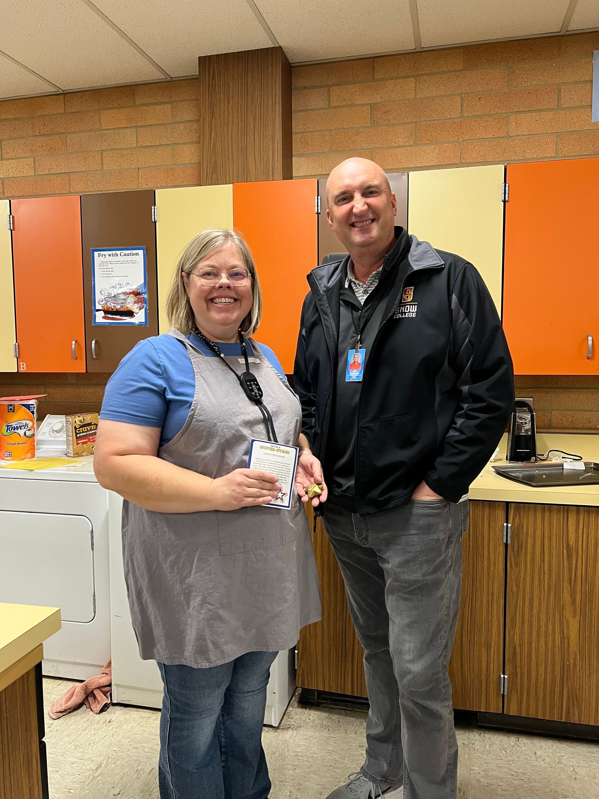 Two people standing in a kitchen with orange cabinets. Woman in apron smiles, man in jacket smiles.