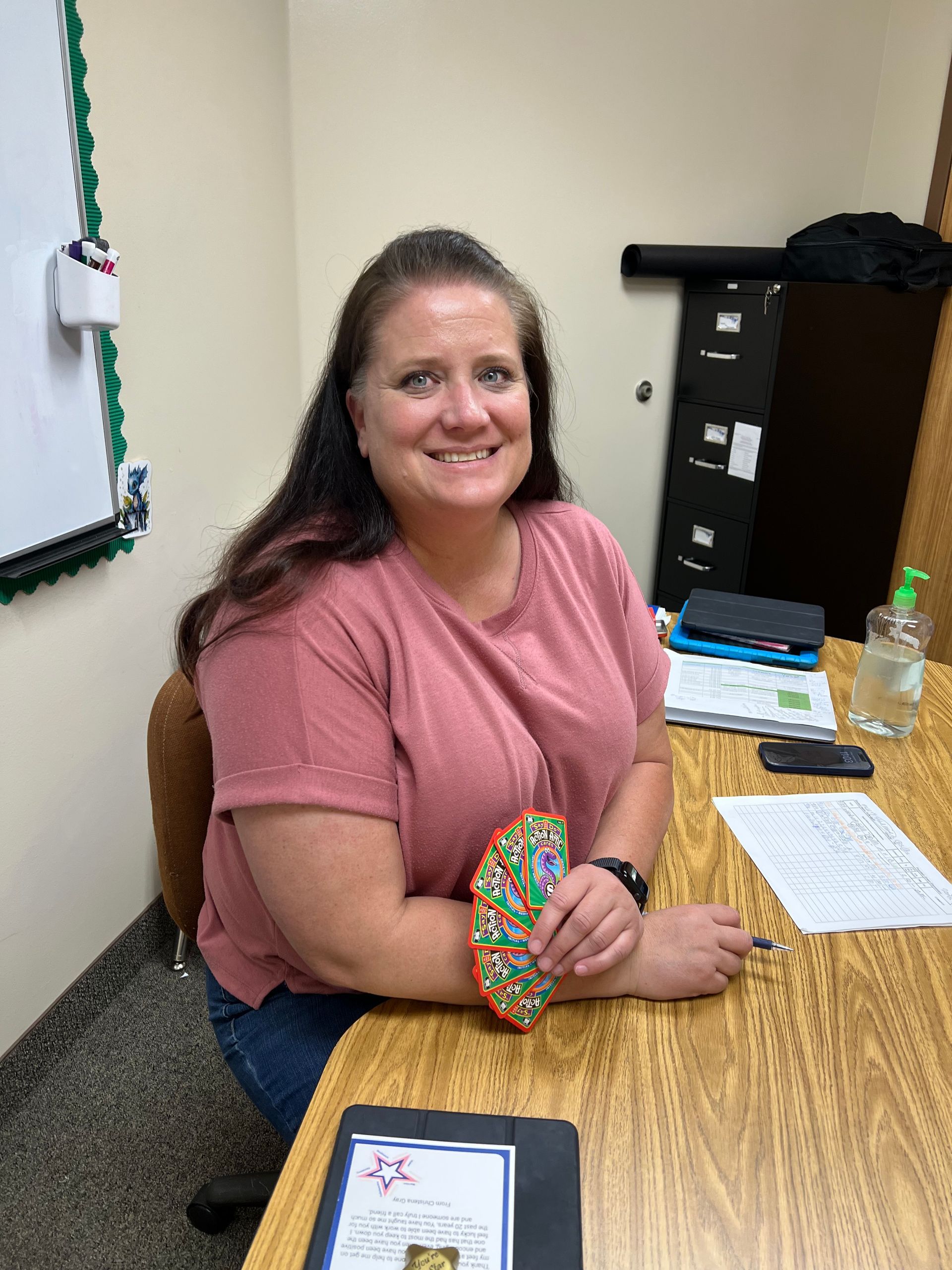 Woman with brown hair smiles, sitting at a desk. She wears a pink shirt and holds a colorful fan.
