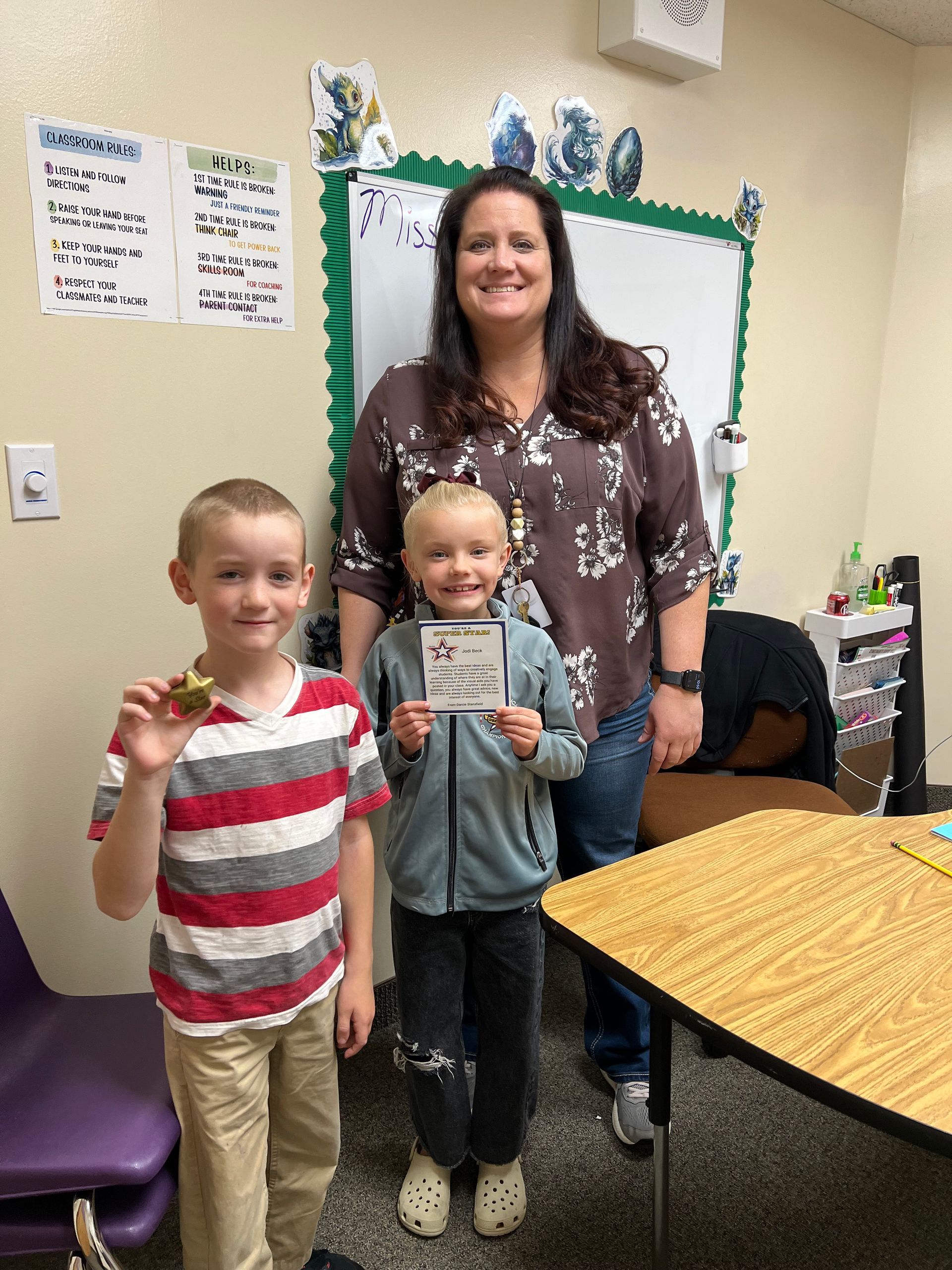 Two children and a woman in a classroom setting. One child holds a coin, the other a card. Woman smiles.
