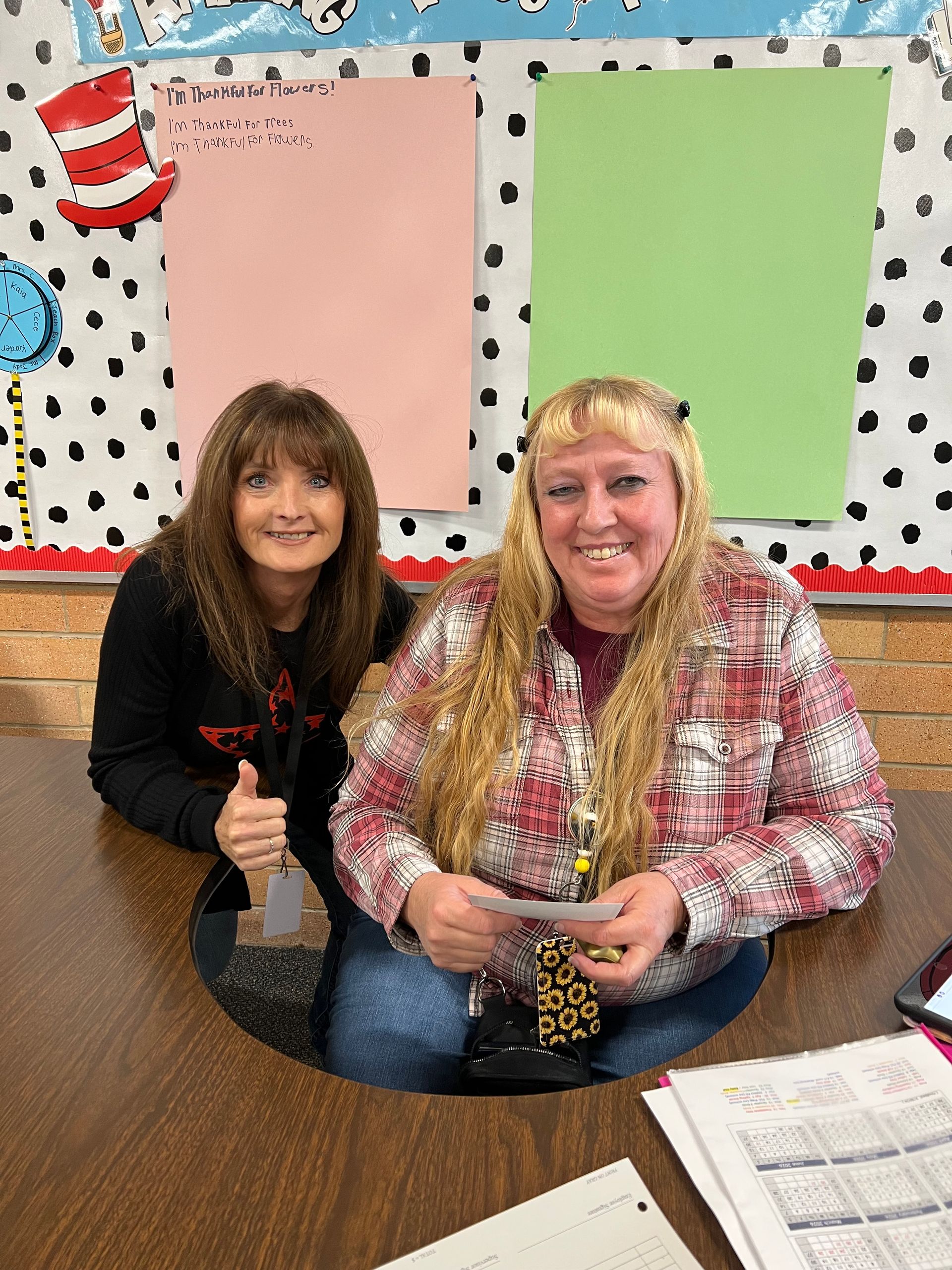 Two women at a desk; one gives a thumbs up. Classroom background with Dr. Seuss decorations.
