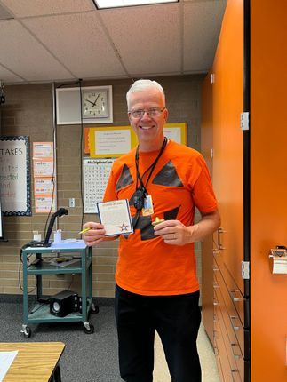 Man in pumpkin shirt holds papers in classroom. Orange and black color scheme.