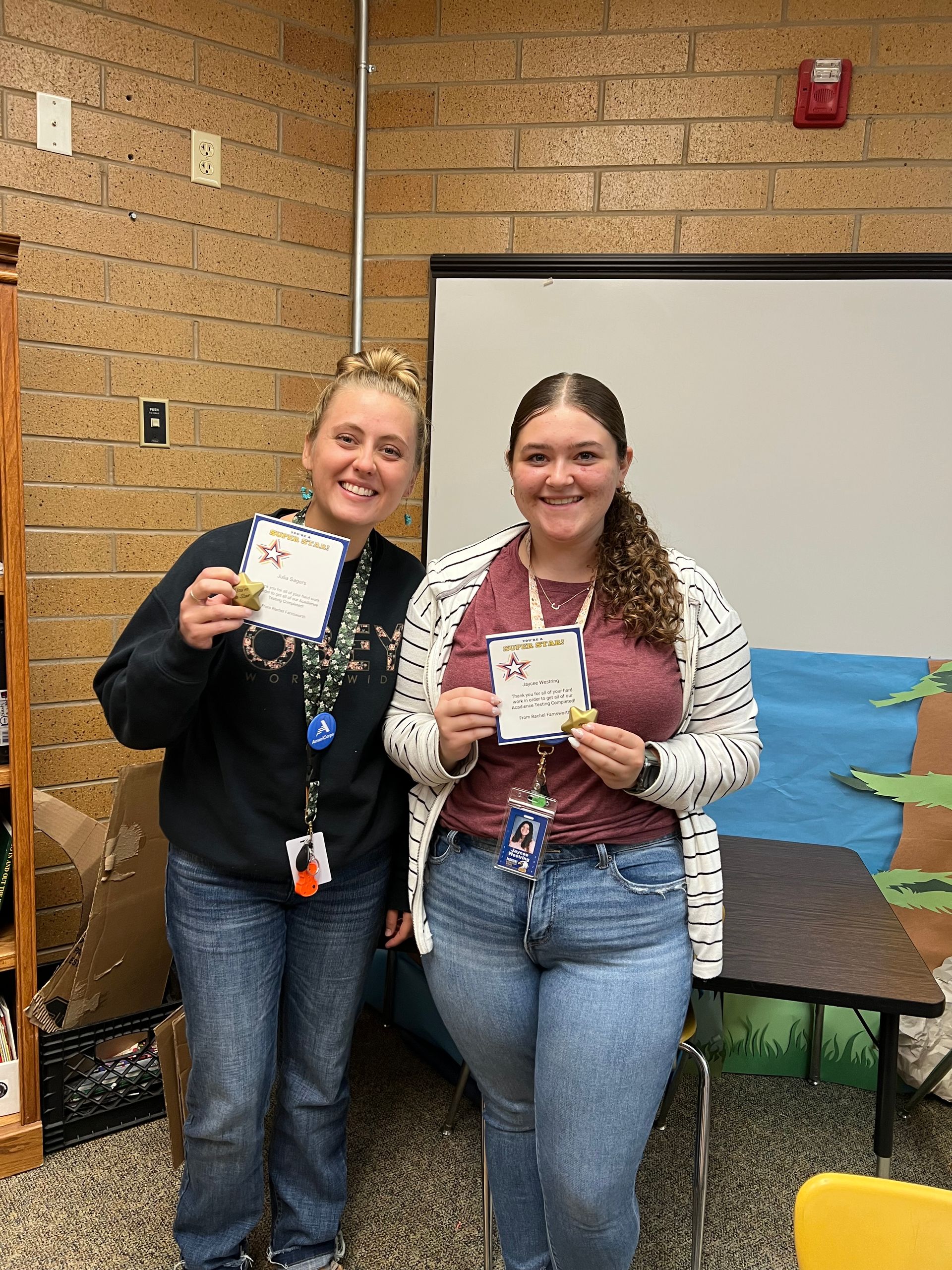 Two women holding cards, smiling, in a classroom. One wears a black sweatshirt and jeans, the other a white cardigan and jeans.