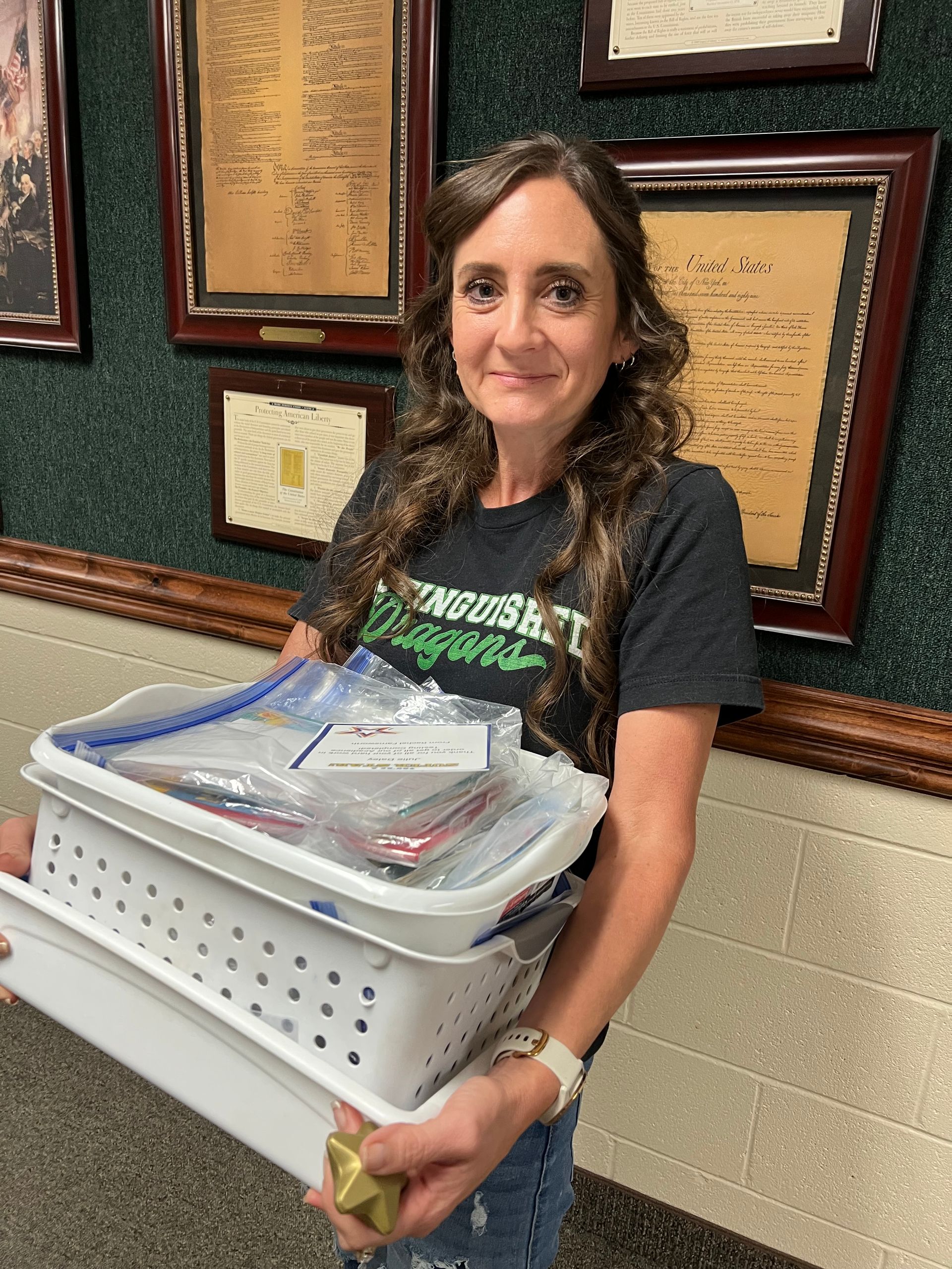 Woman holds a white basket filled with supplies, smiling in front of framed documents.