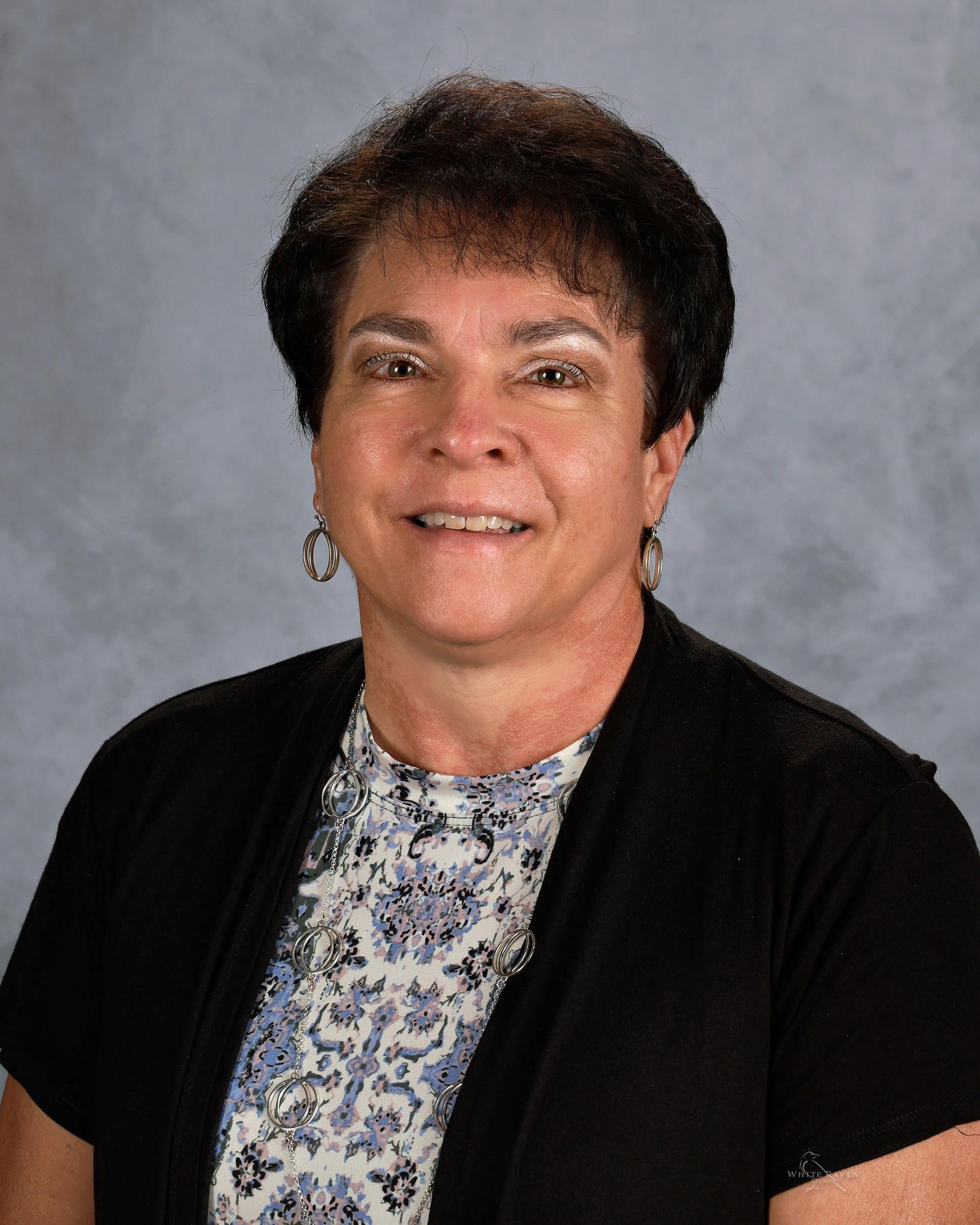 Woman with short dark hair smiling, wearing a floral top, black sweater, against a gray background.
