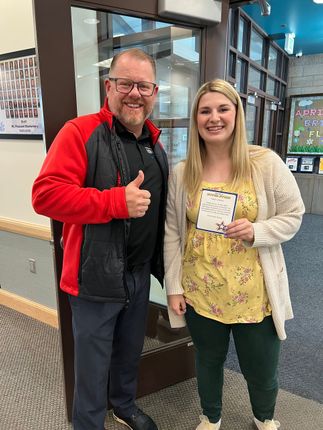 Two people smiling indoors, one giving a thumbs-up and the other holding a certificate.