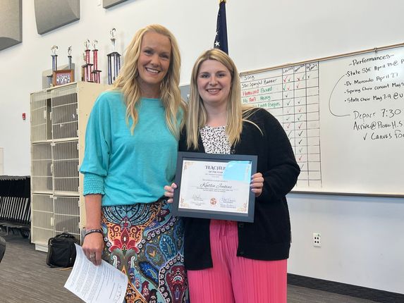 Two women smiling in a classroom, holding a certificate in front of a whiteboard.