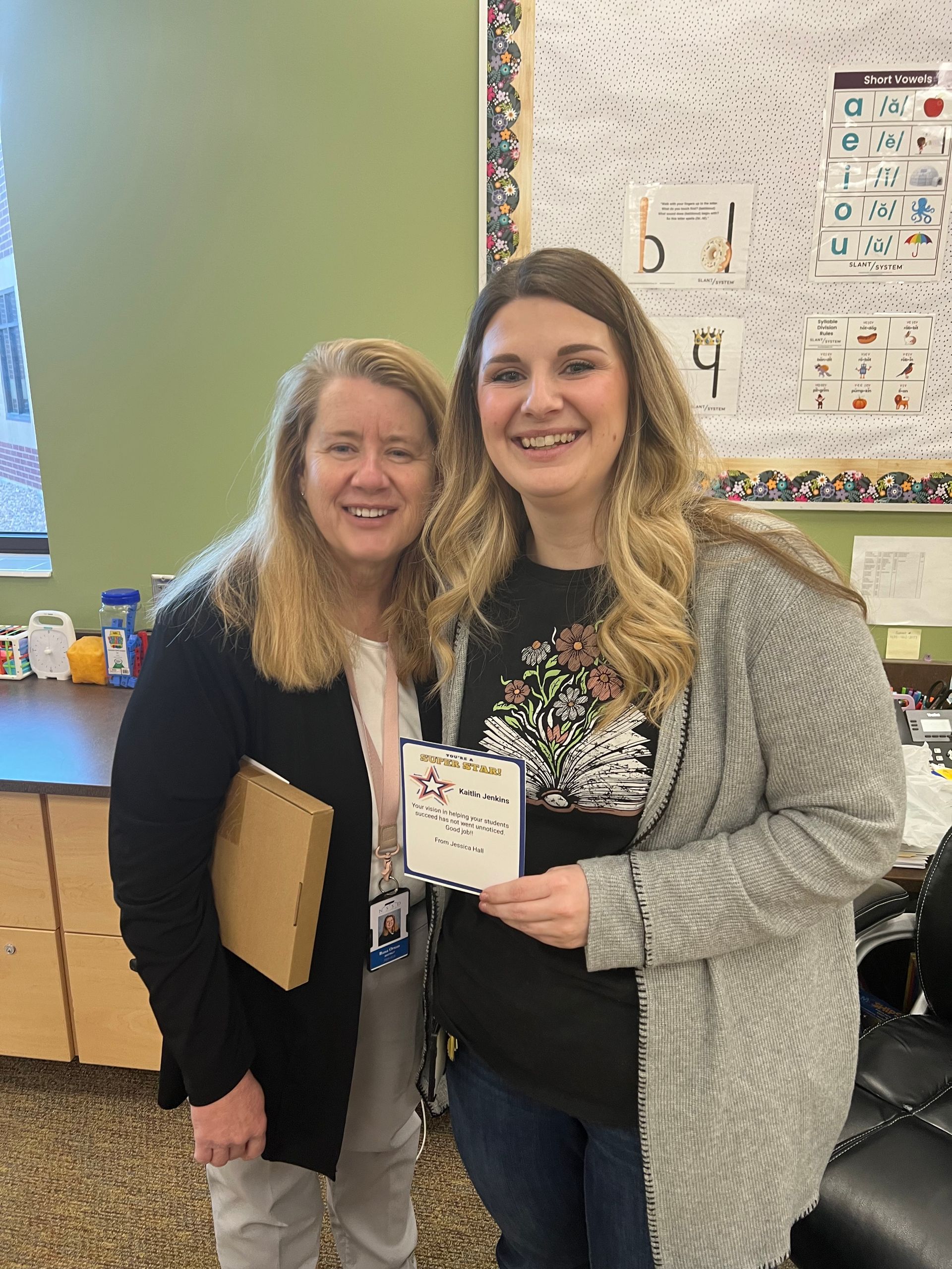 Two smiling women stand in a classroom, one holding a gift box and the other holding a small card.