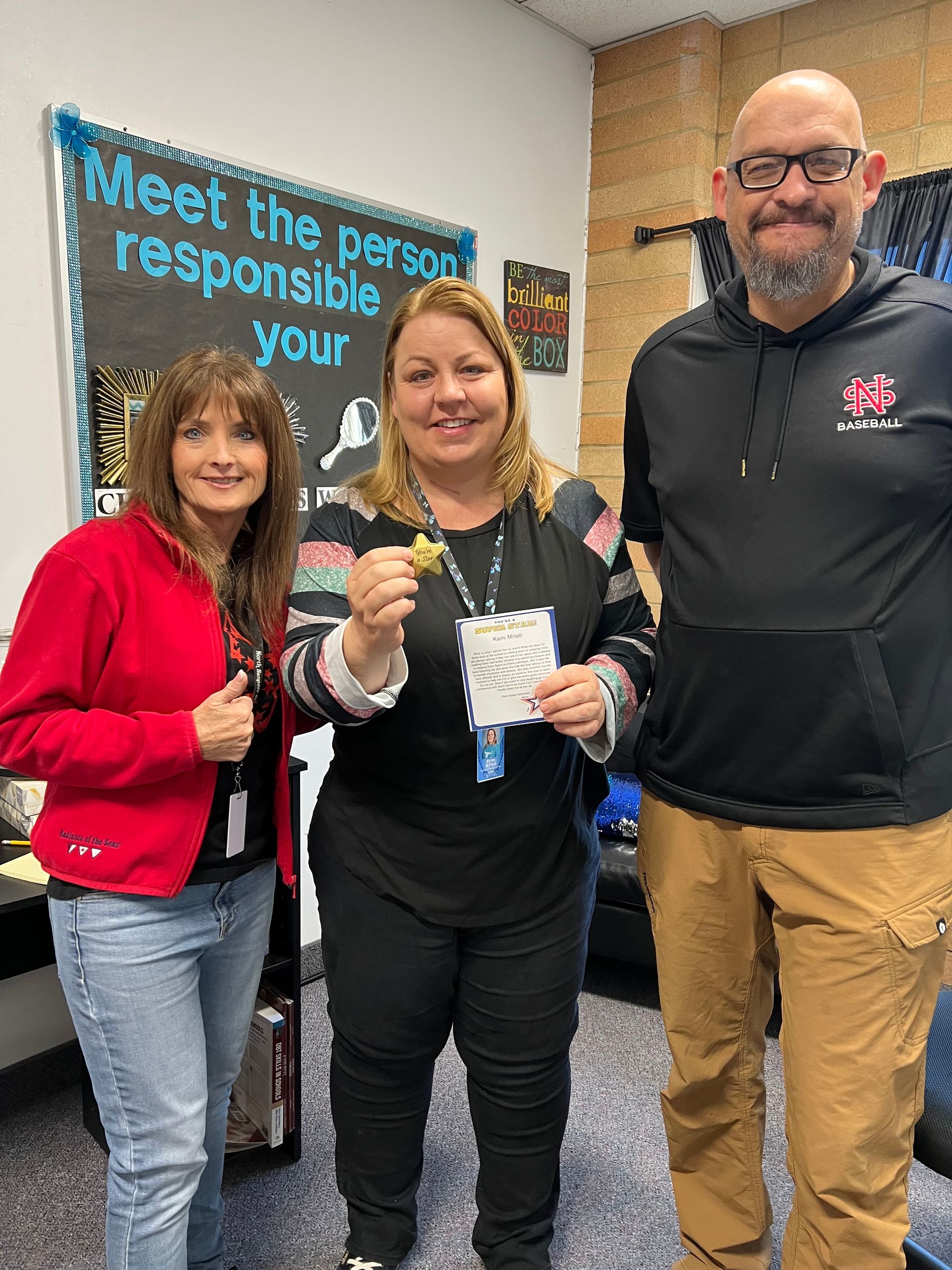 Three people in a classroom. Woman in the center holds a star and certificate. Others smile and give thumbs up.