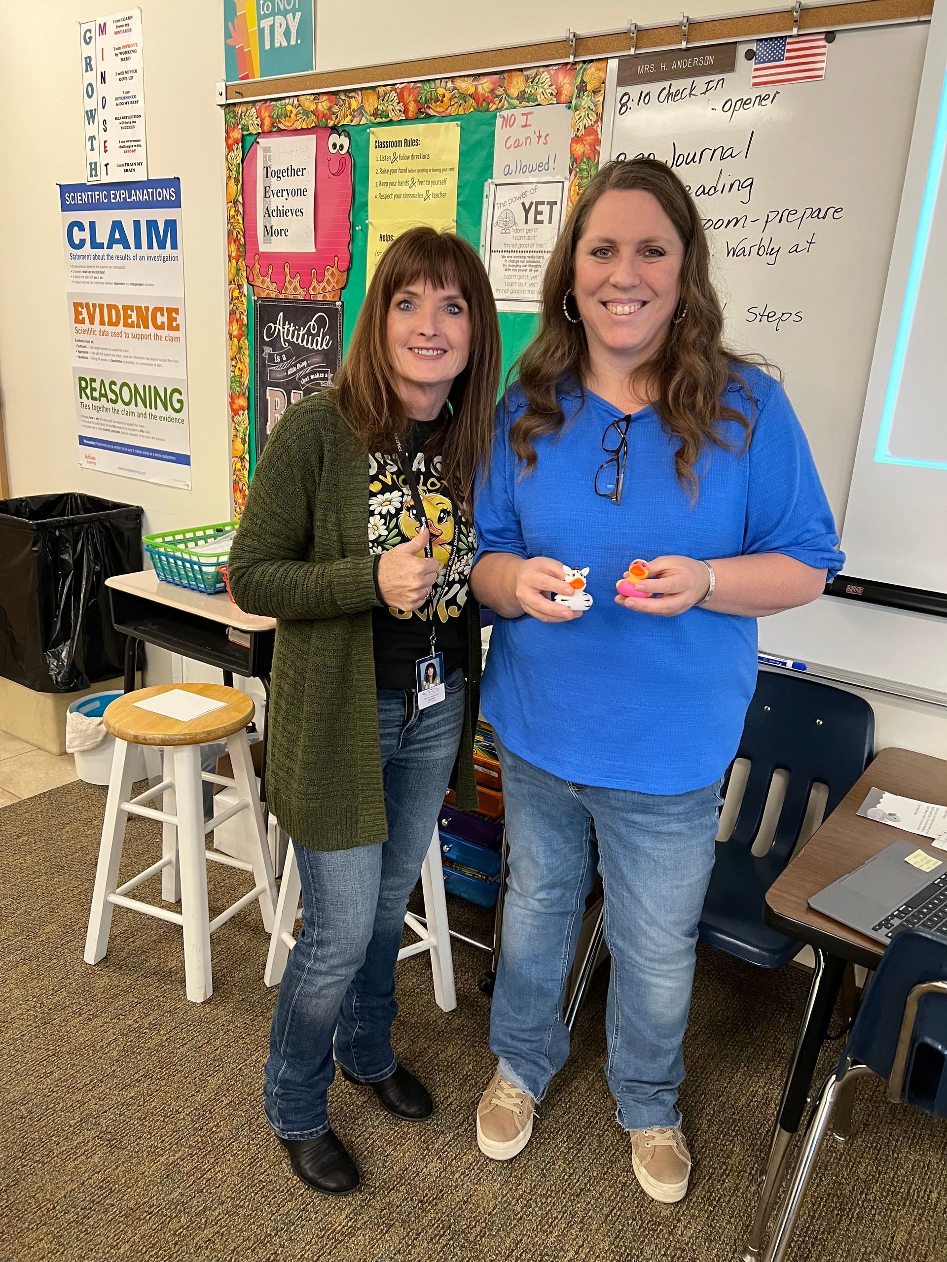 Two women are standing next to each other in a classroom holding cupcakes.